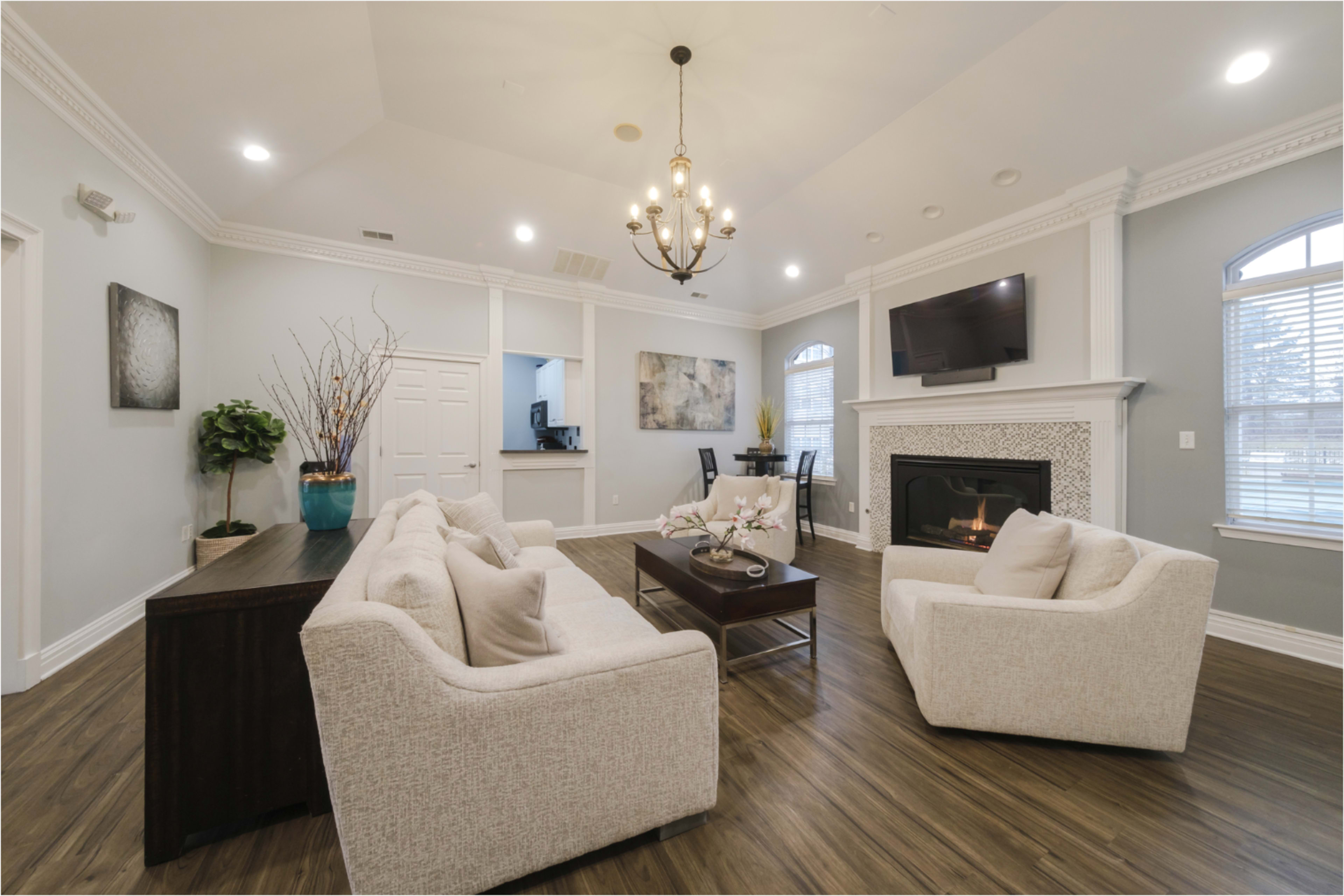 The image shows a spacious living room with two white sofas, a dark coffee table, and a fireplace, featuring a chandelier and large windows.