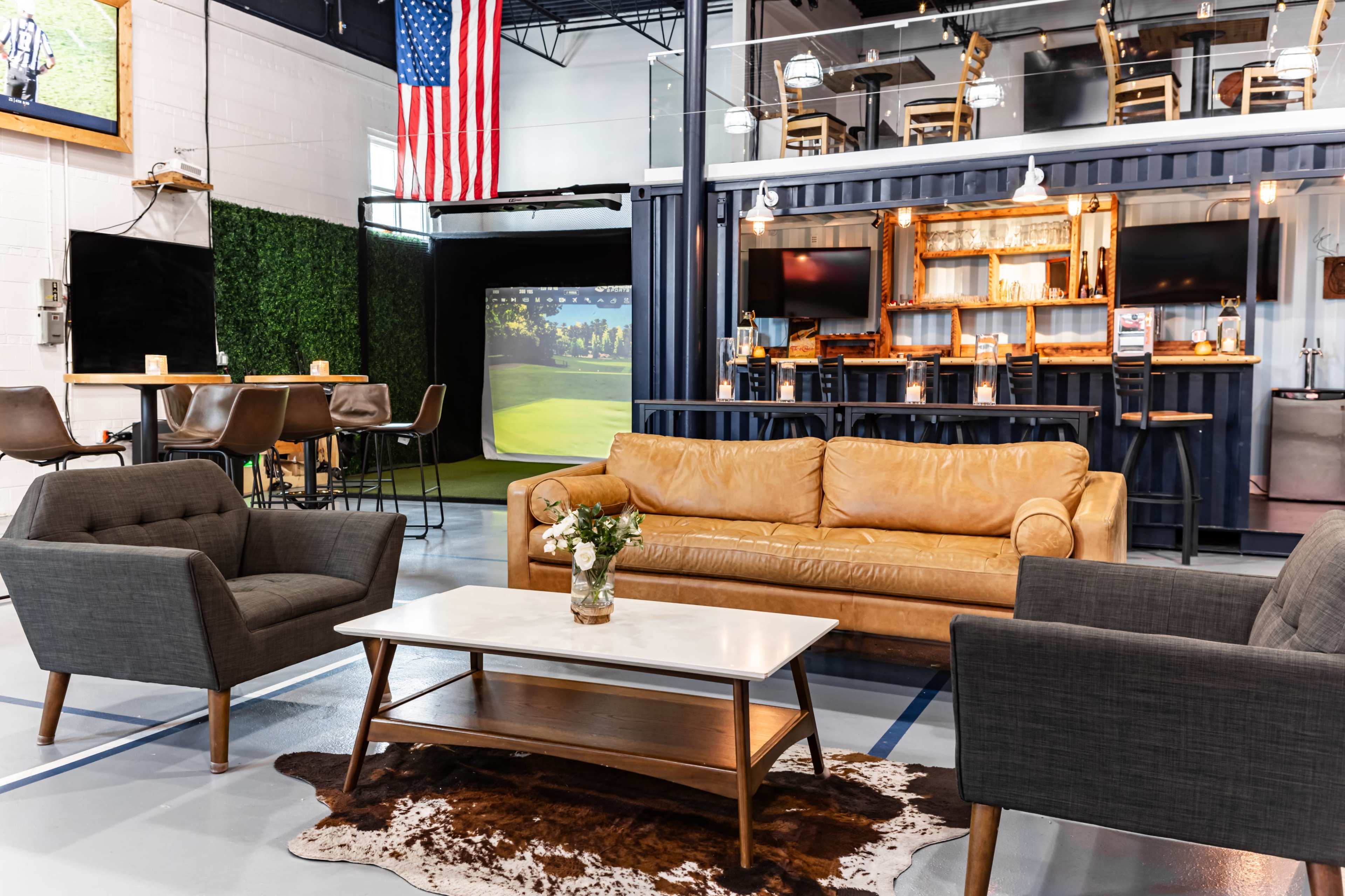 A modern indoor lounge area featuring a brown leather sofa, two armchairs, a coffee table, and a golf simulation setup in the background, all under an American flag display.