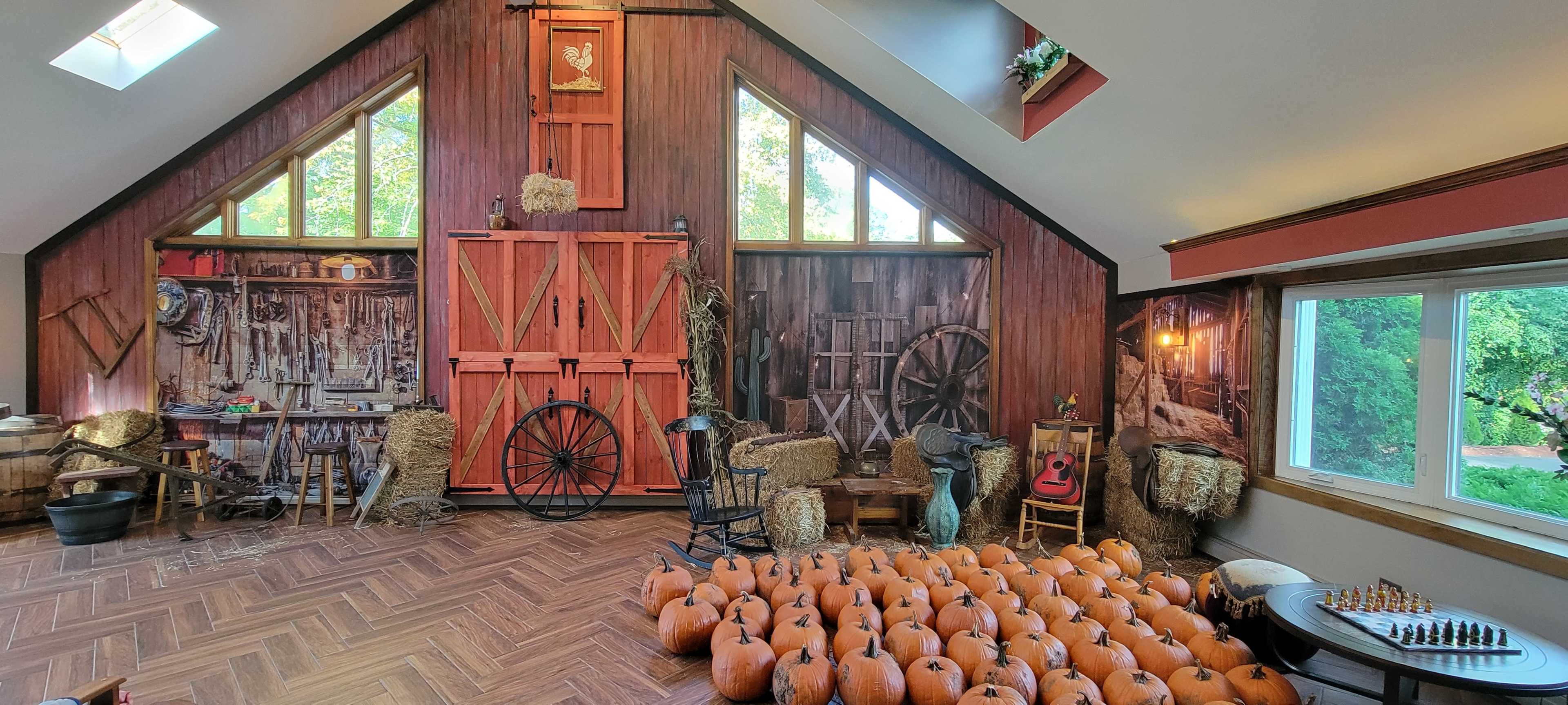 The interior of a rustic barn features a wall adorned with tools and decorations, while a large collection of pumpkins is arranged in front.