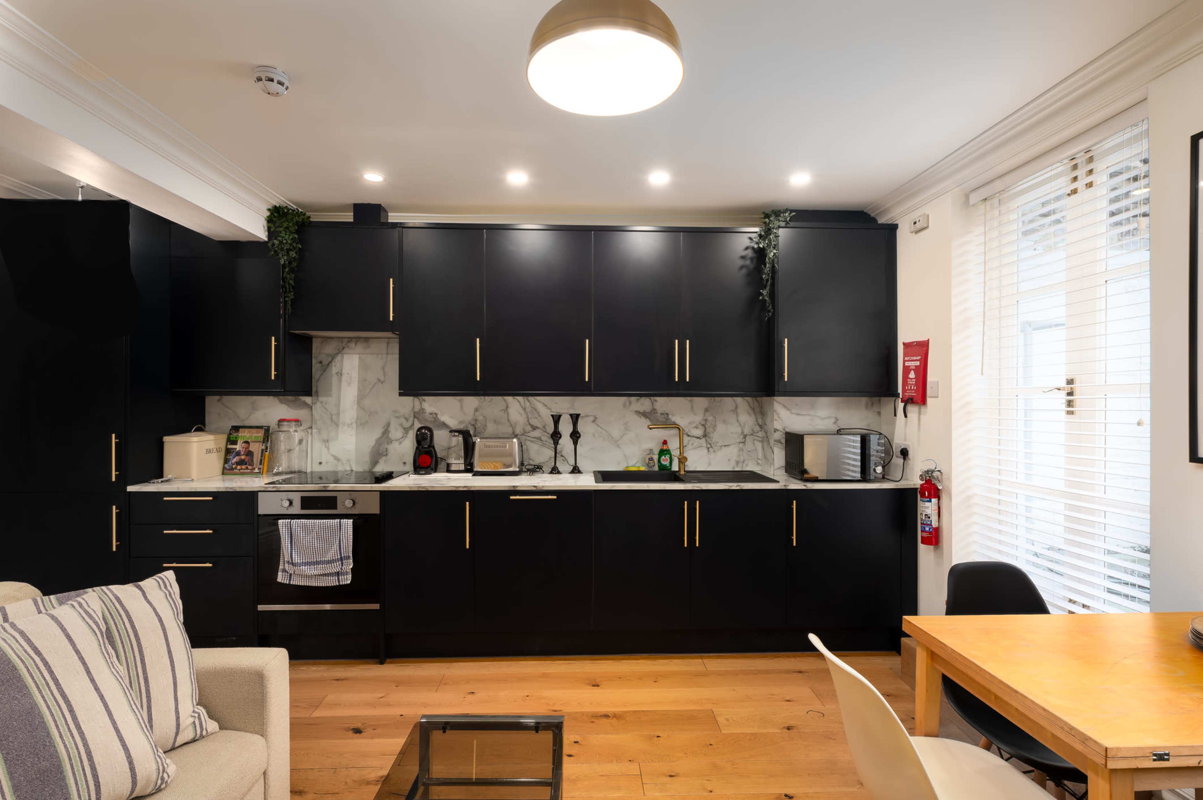 The image shows a modern kitchen with dark cabinets, marble countertops, and stainless steel appliances, adjacent to a living area.