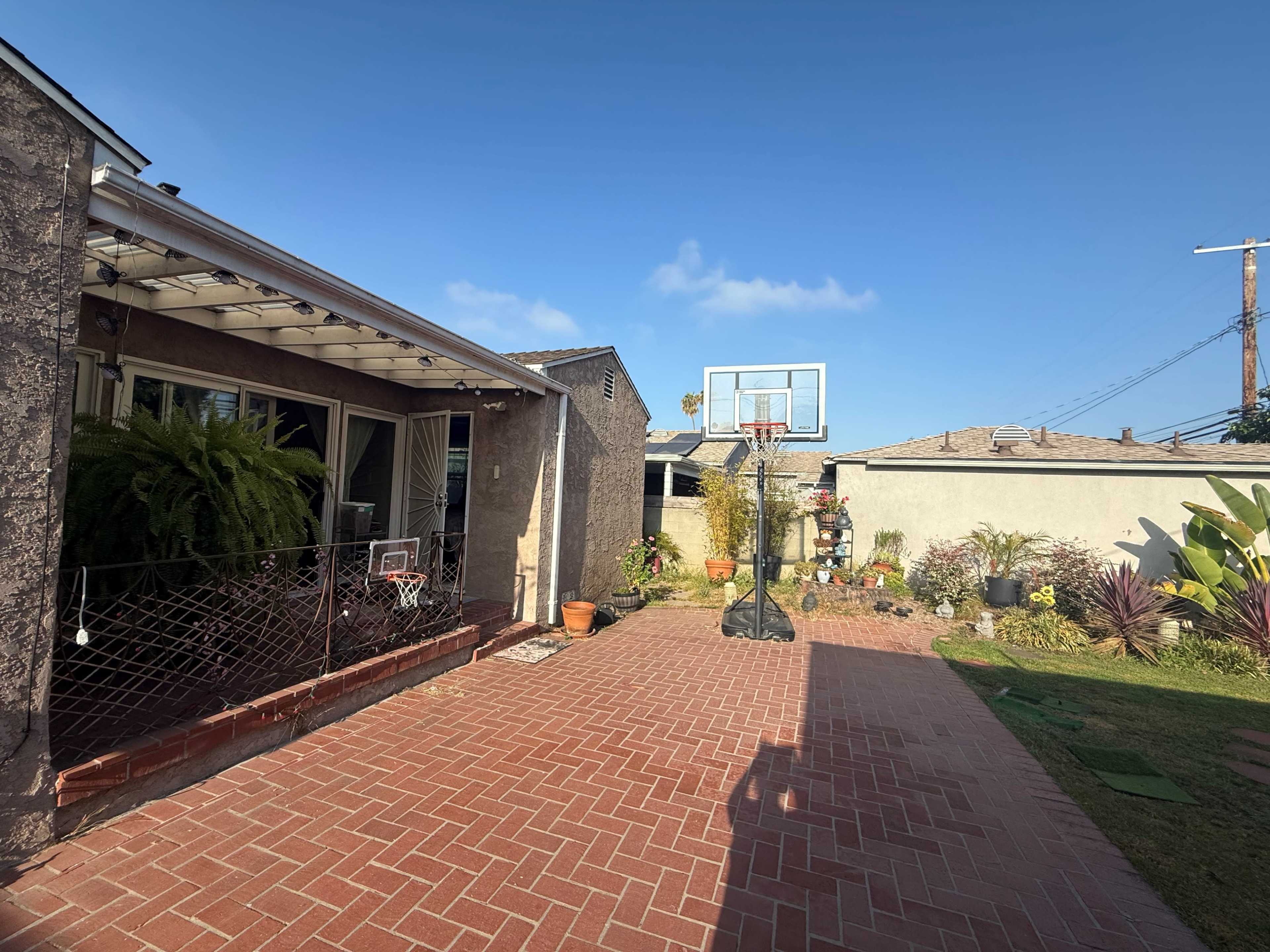 The image shows a backyard area with a brick patio, a basketball hoop, and various plants and landscaping features.