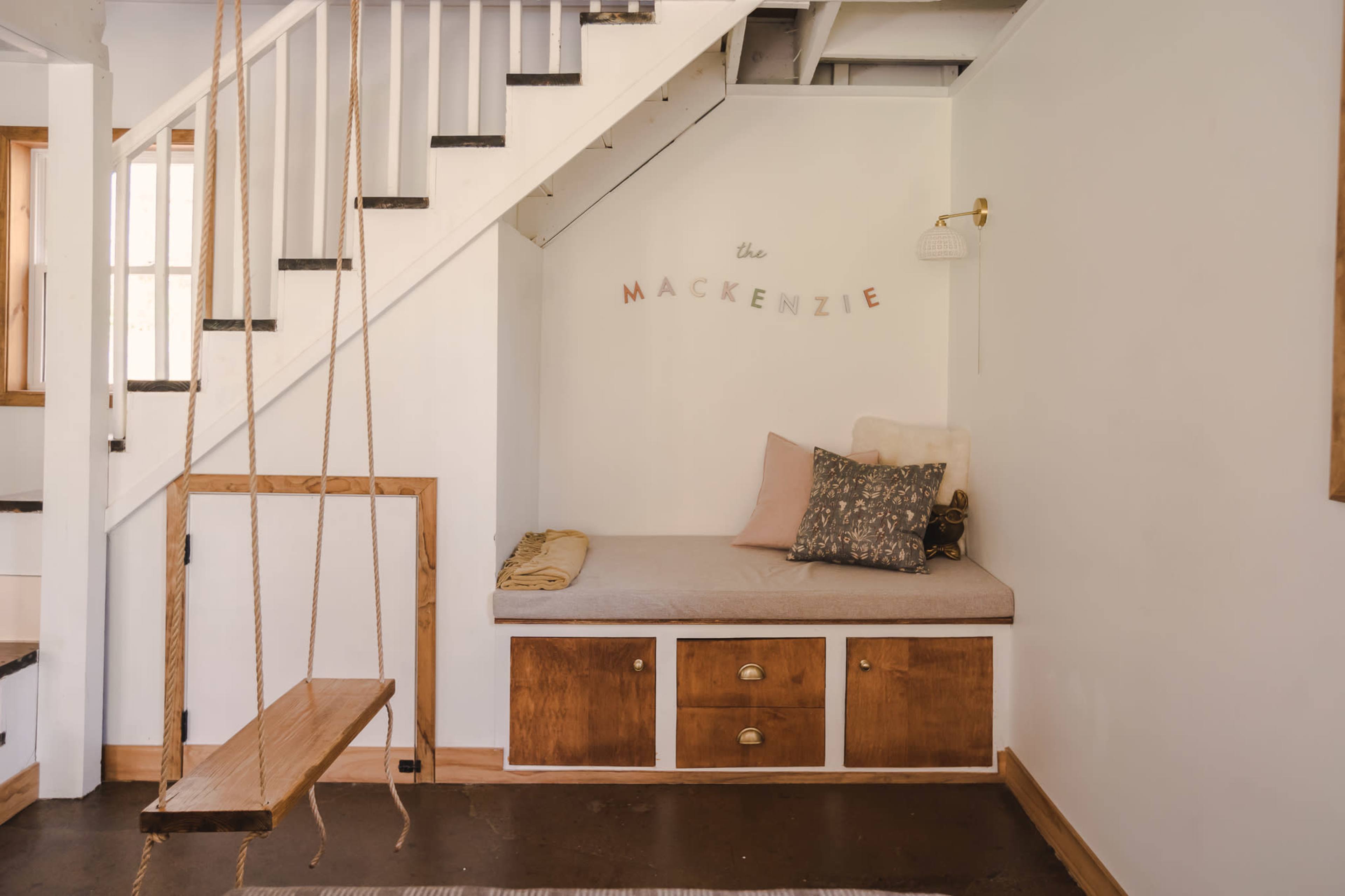 The image shows a cozy nook with a cushioned bench below a staircase, featuring a swing seat and decorative wall lettering.