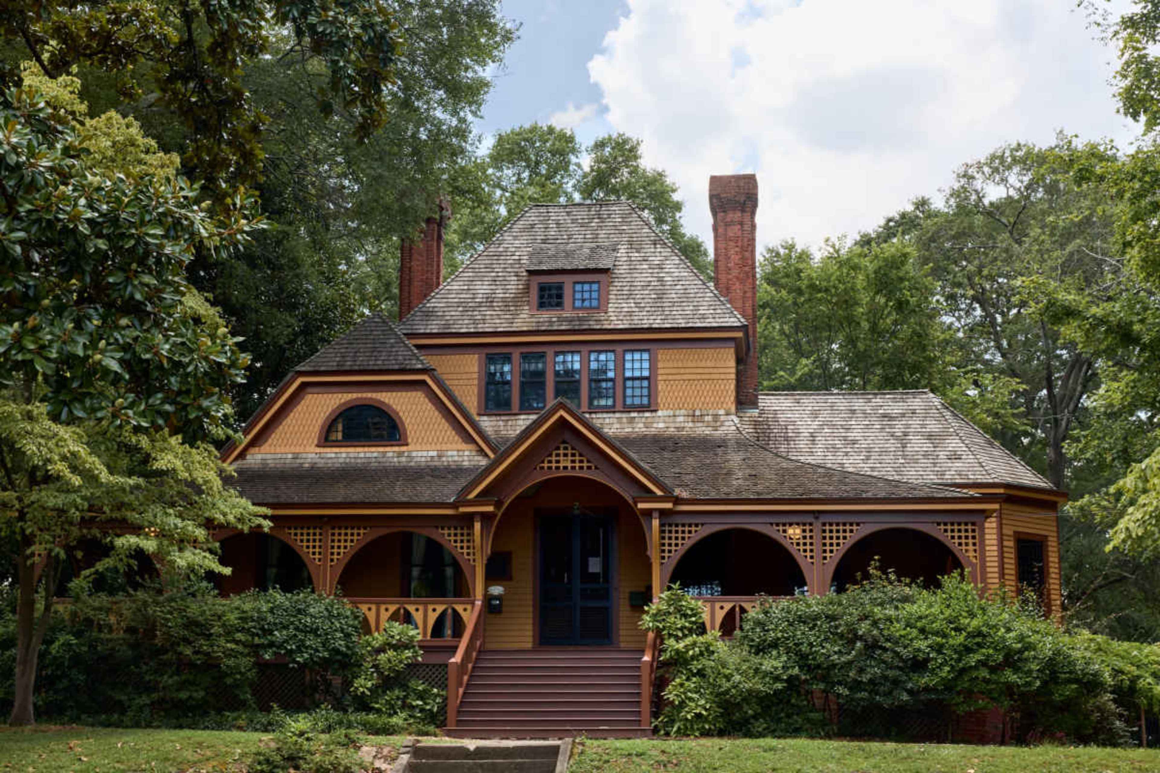 The image shows a large, three-story Victorian-style house with a gable roof, multiple chimneys, and a wraparound porch surrounded by trees and shrubbery.