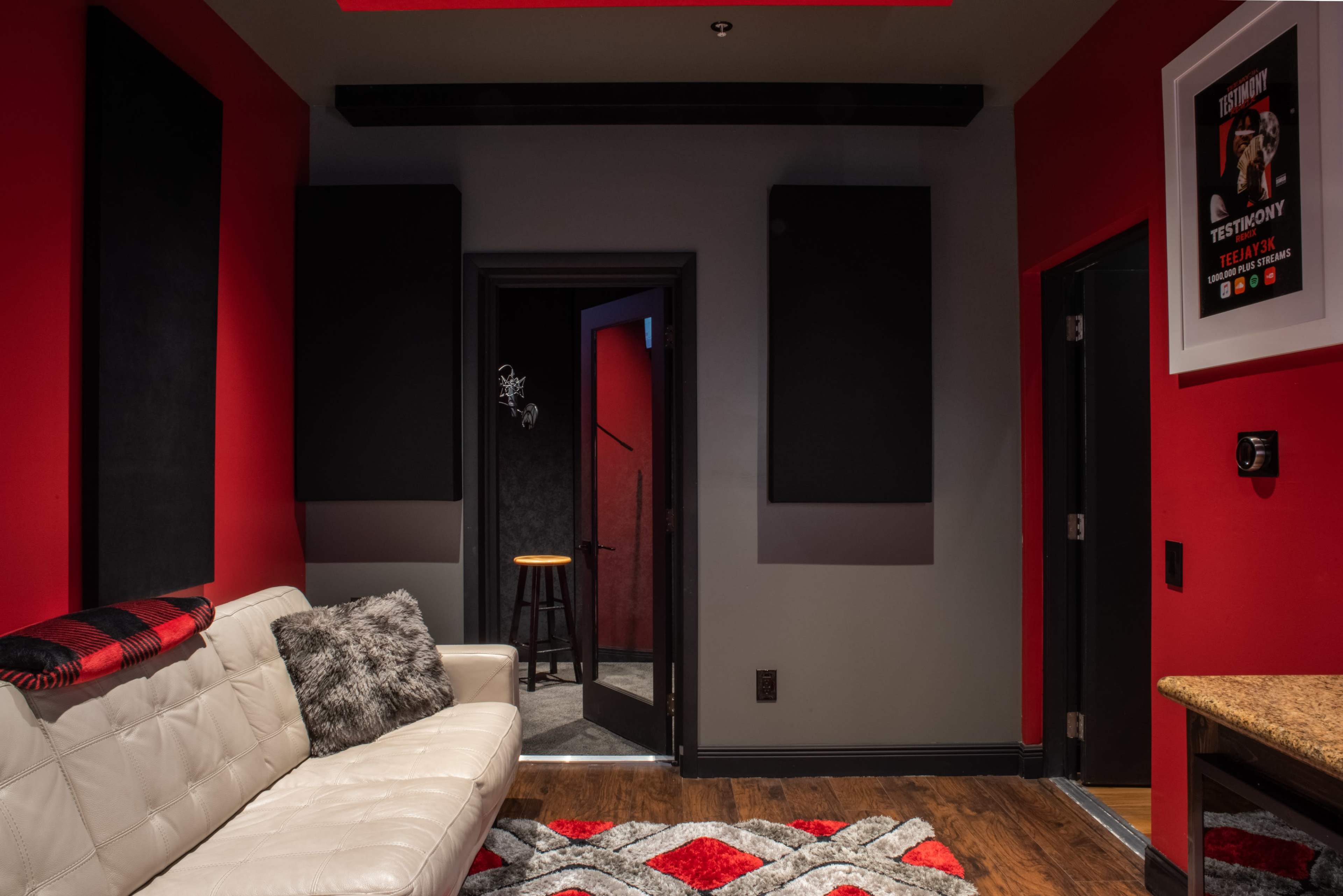 A modern room with a white leather sofa, a red and gray rug, and a doorway leading to a dark space, accented by black soundproofing panels on the walls.