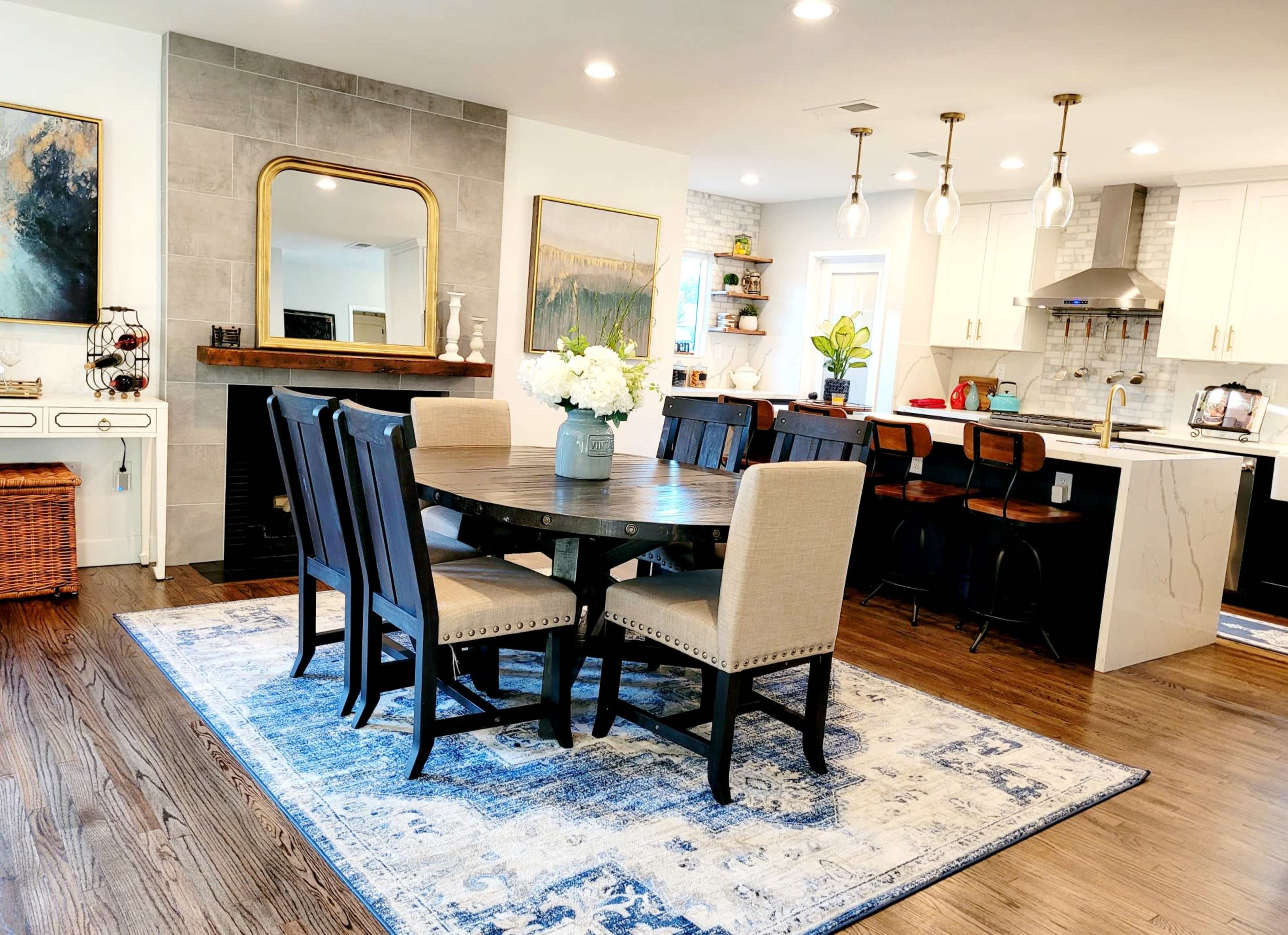 A modern kitchen and dining area with a wooden dining table, upholstered chairs, and a kitchen counter with bar stools, all arranged around a decorative rug.
