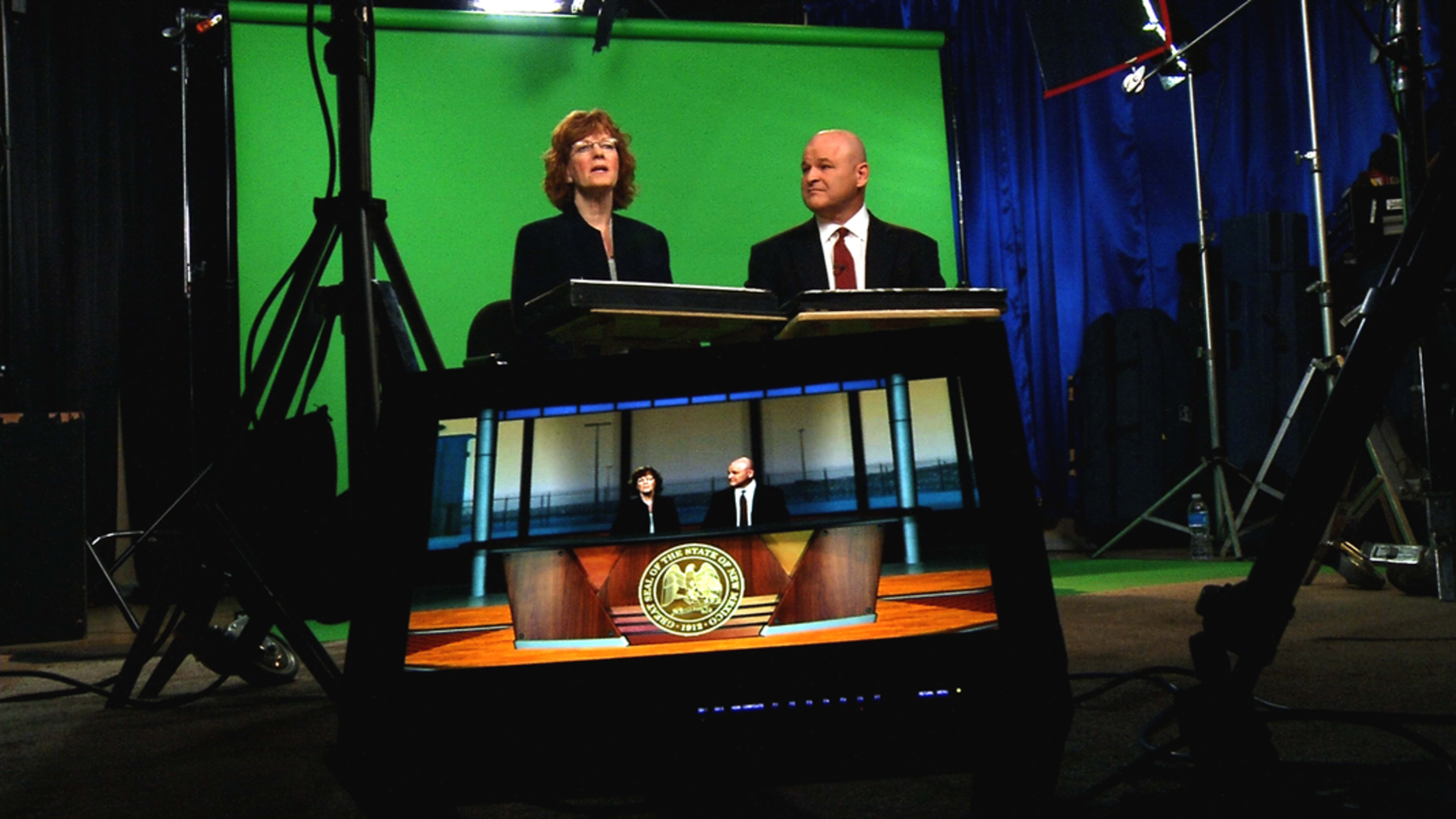 Two presenters seated in front of a green screen, with a monitor displaying a virtual news set in the foreground.