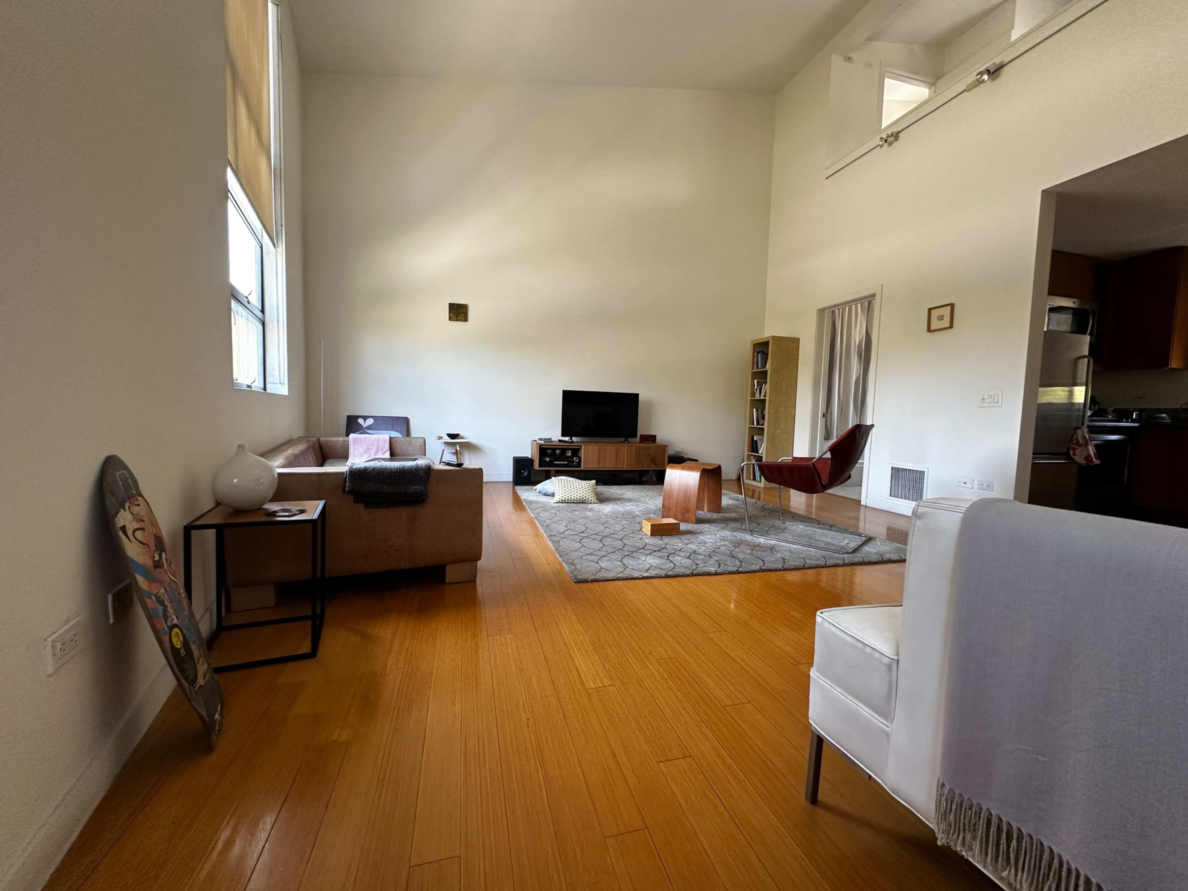 The image shows a spacious living room with wooden flooring, featuring a television on a stand, a sectional couch, a rug, and various furniture pieces.