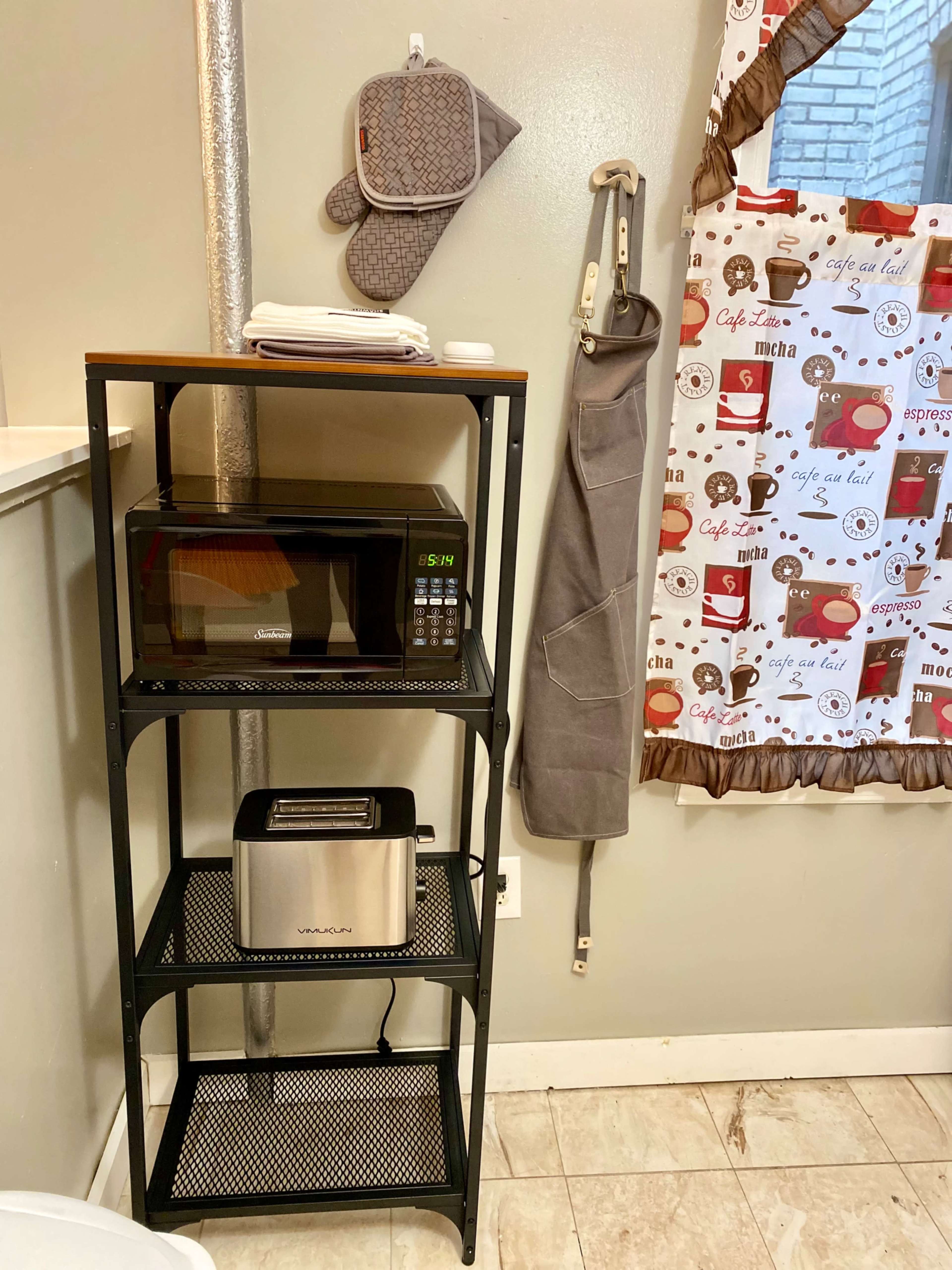 The image shows a three-tier kitchen shelving unit with a microwave, toaster, and various kitchen towels, positioned next to a grey apron and an oven mitt hanging on the wall.