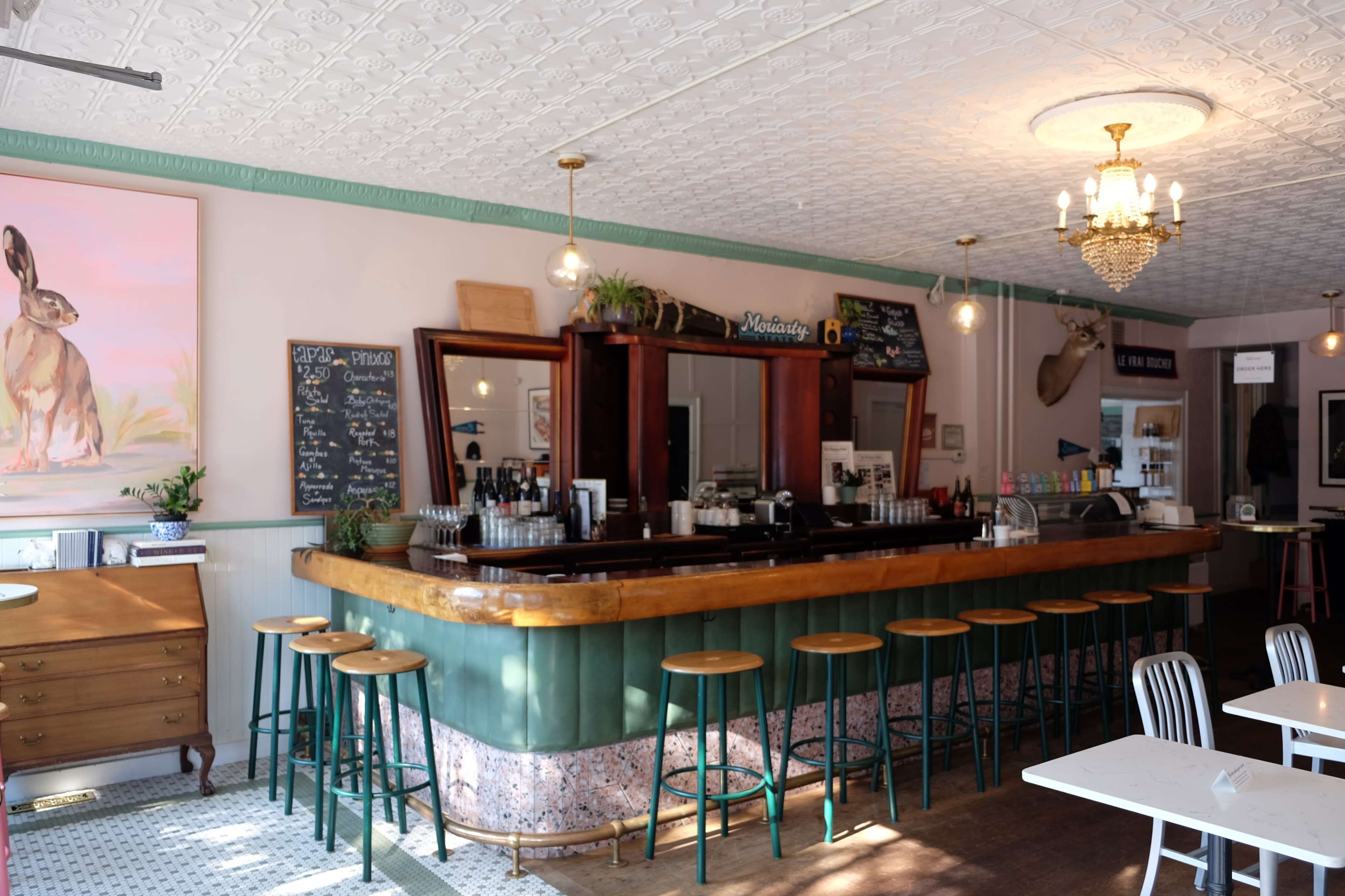 A bar area with wooden stools, a decorative counter, and a wall adorned with a painting and a chalkboard menu.