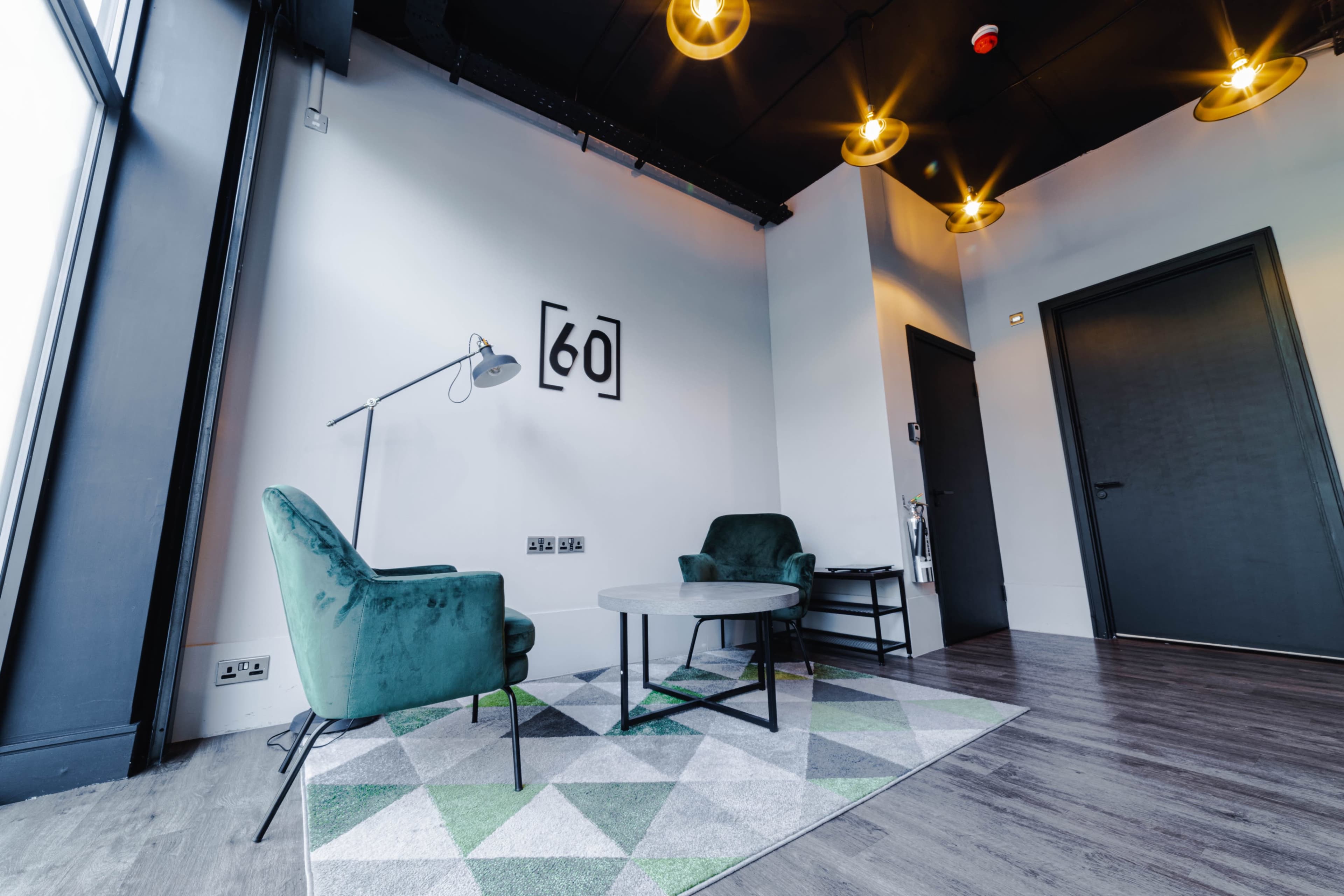 The image shows a modern sitting area with two green chairs, a round coffee table, and a geometric rug under pendant lights and a wall featuring the number "[60]."