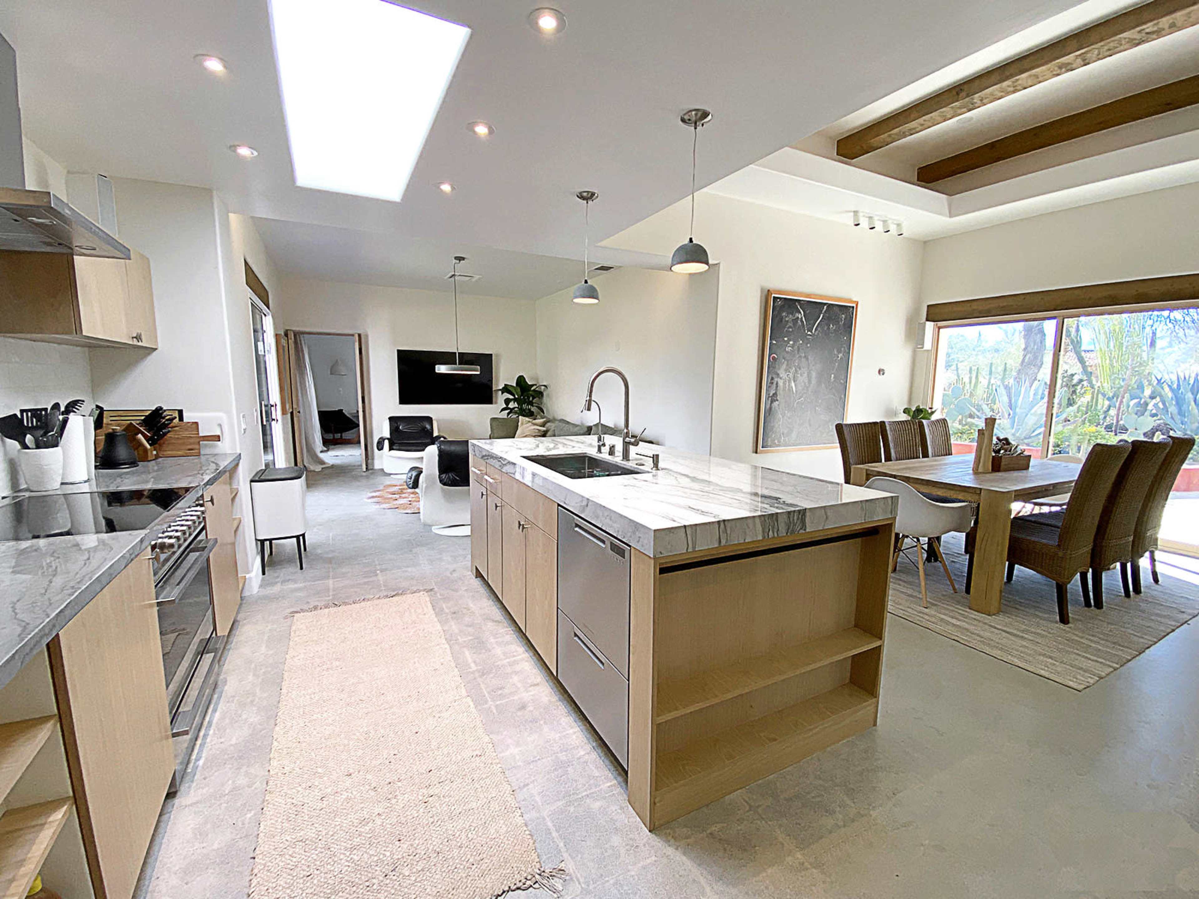 The image shows a modern kitchen with a marble island and countertops, seamlessly connected to a dining area with a large window and natural light.