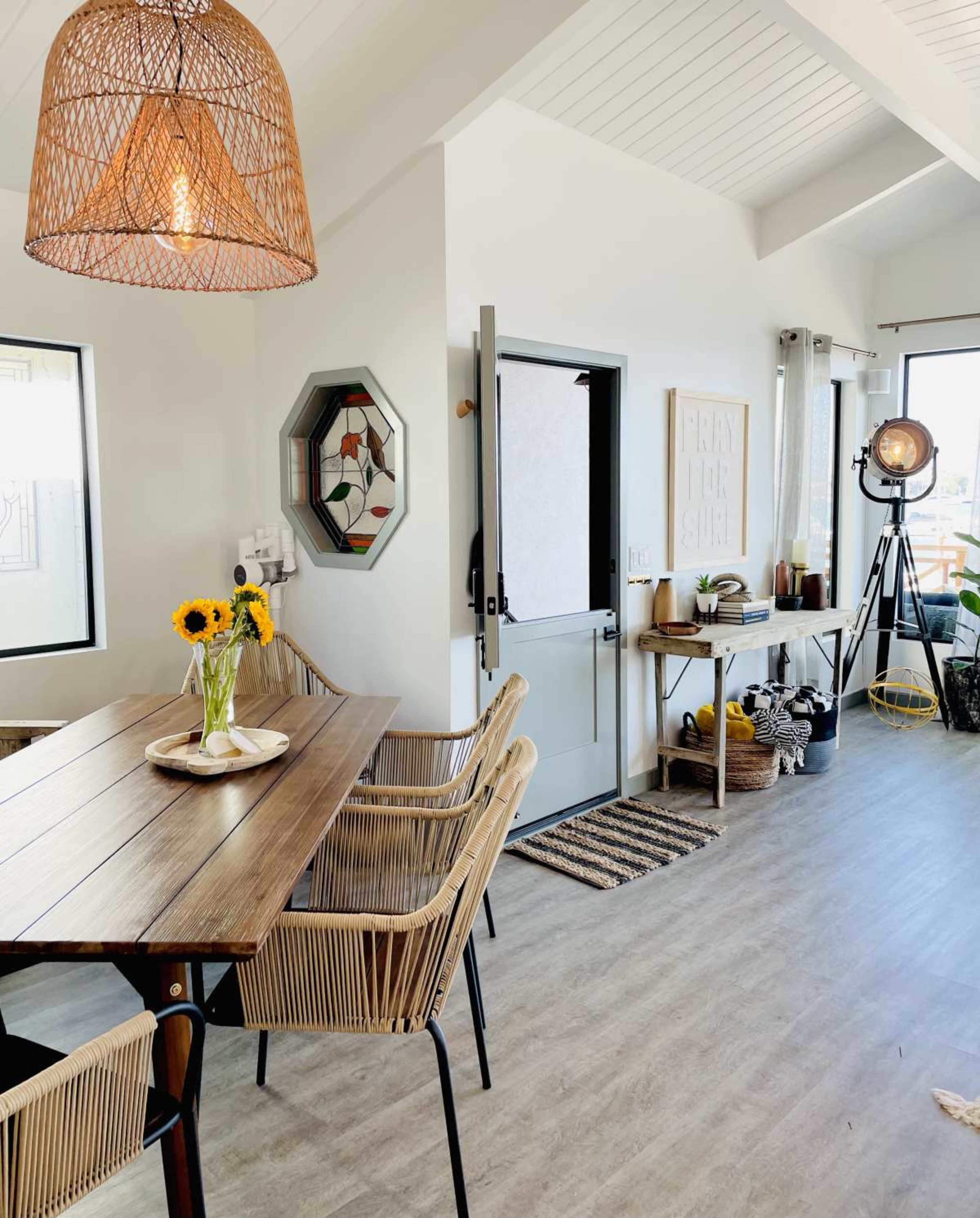 A dining area features a wooden table surrounded by chairs, with a door leading to the outside and a large light fixture overhead.