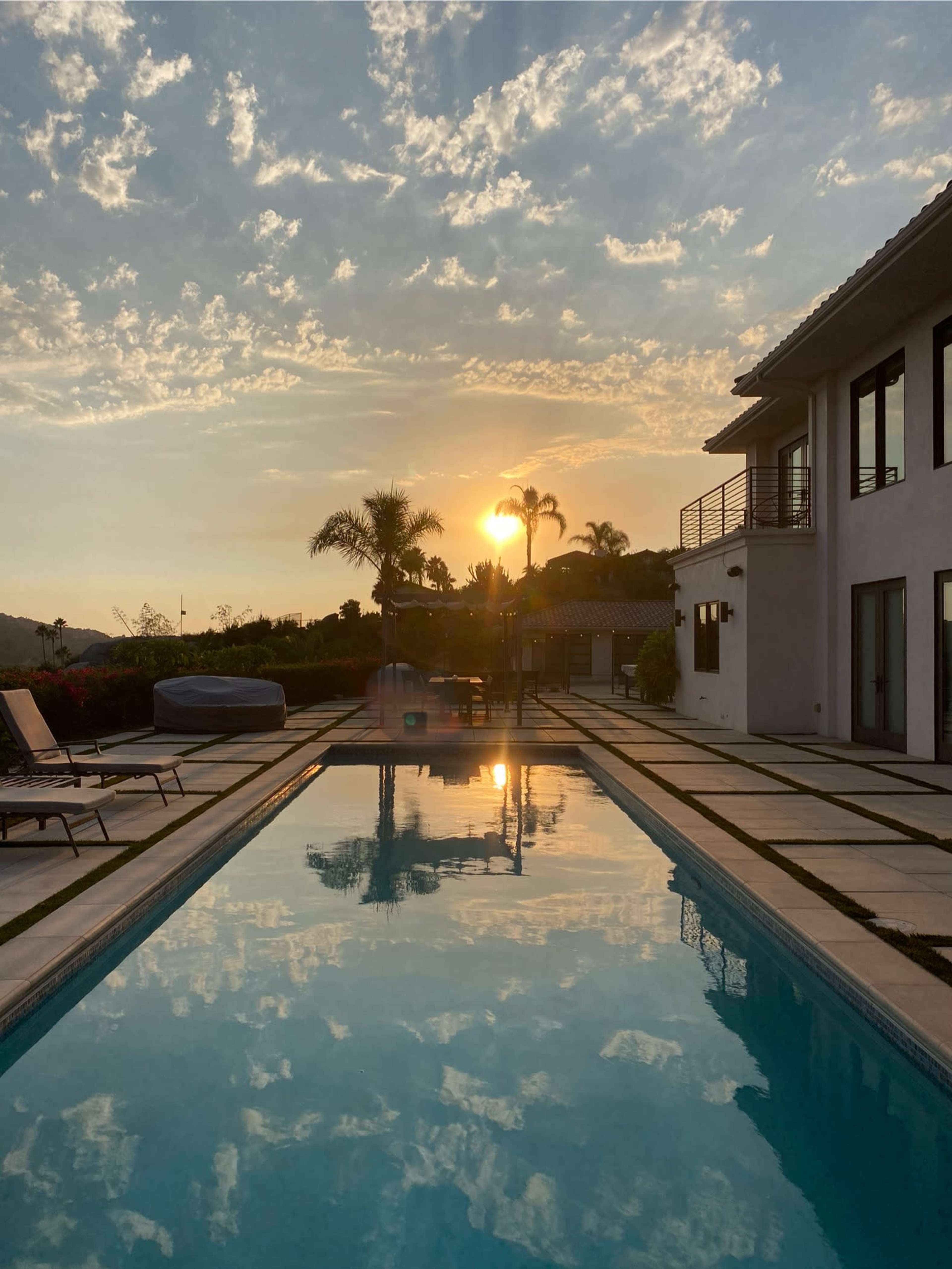 The image shows a swimming pool reflecting a sunset surrounded by palm trees and a modern house.