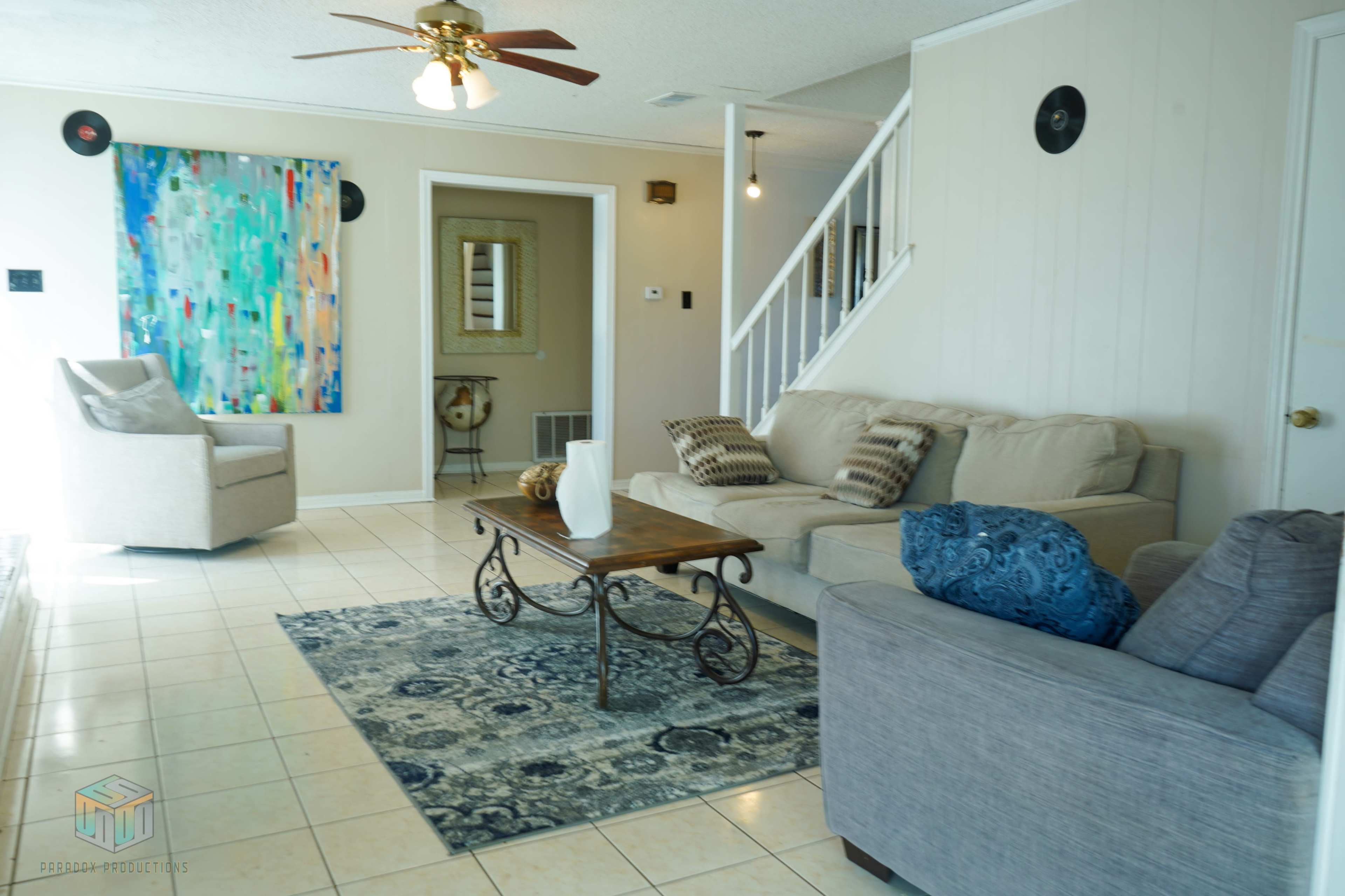 A living room with a beige sofa, a gray armchair, a wooden coffee table, and an area rug, complemented by colorful artwork on the wall and a ceiling fan above.