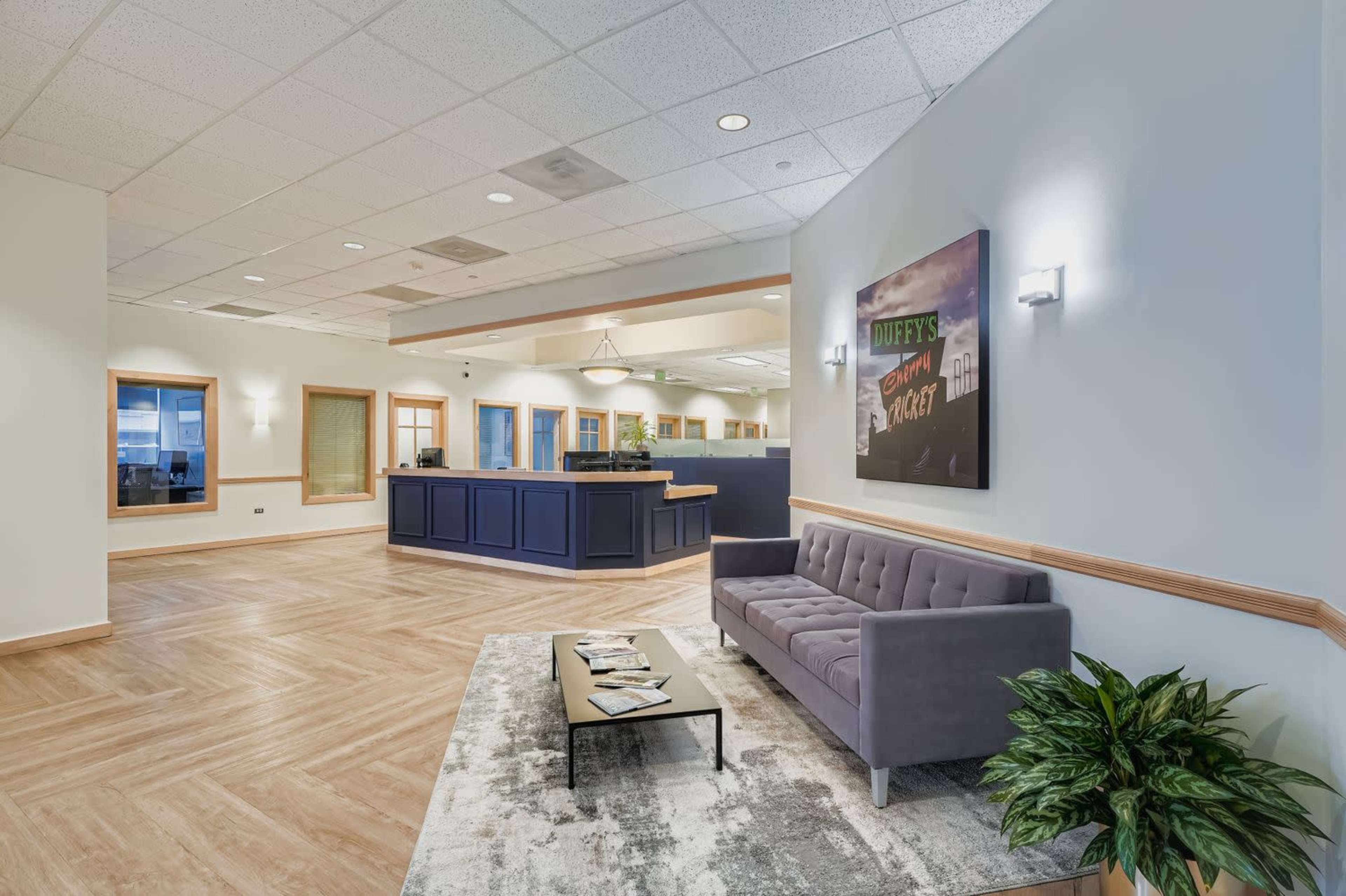 The image shows a waiting area with a gray couch, a coffee table, and a reception desk in a well-lit office space.