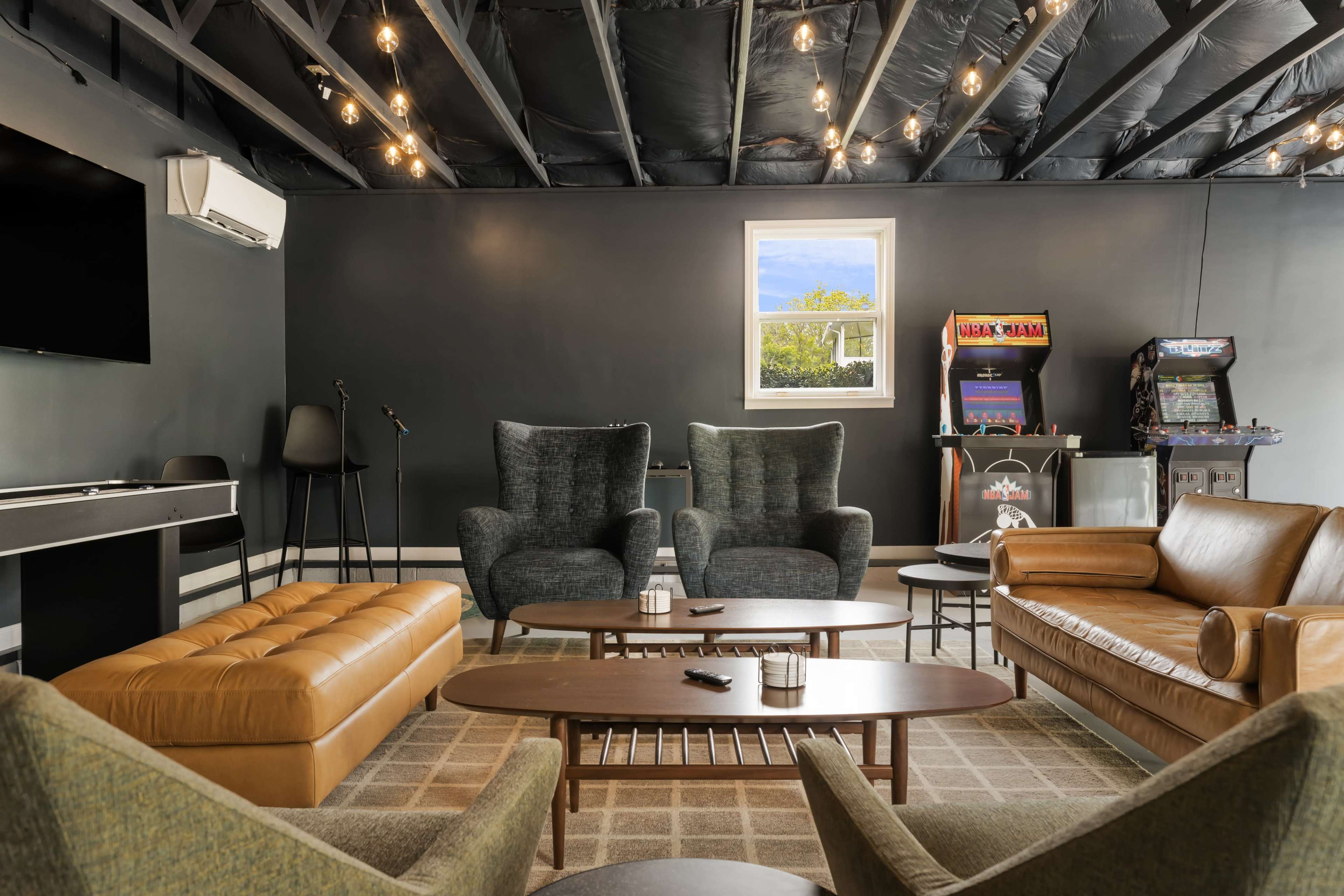 The image features a cozy lounge area with multiple seating options, including two armchairs, a leather sofa, and a tufted bench, arranged around a coffee table, along with an arcade machine and a television mounted on the wall.