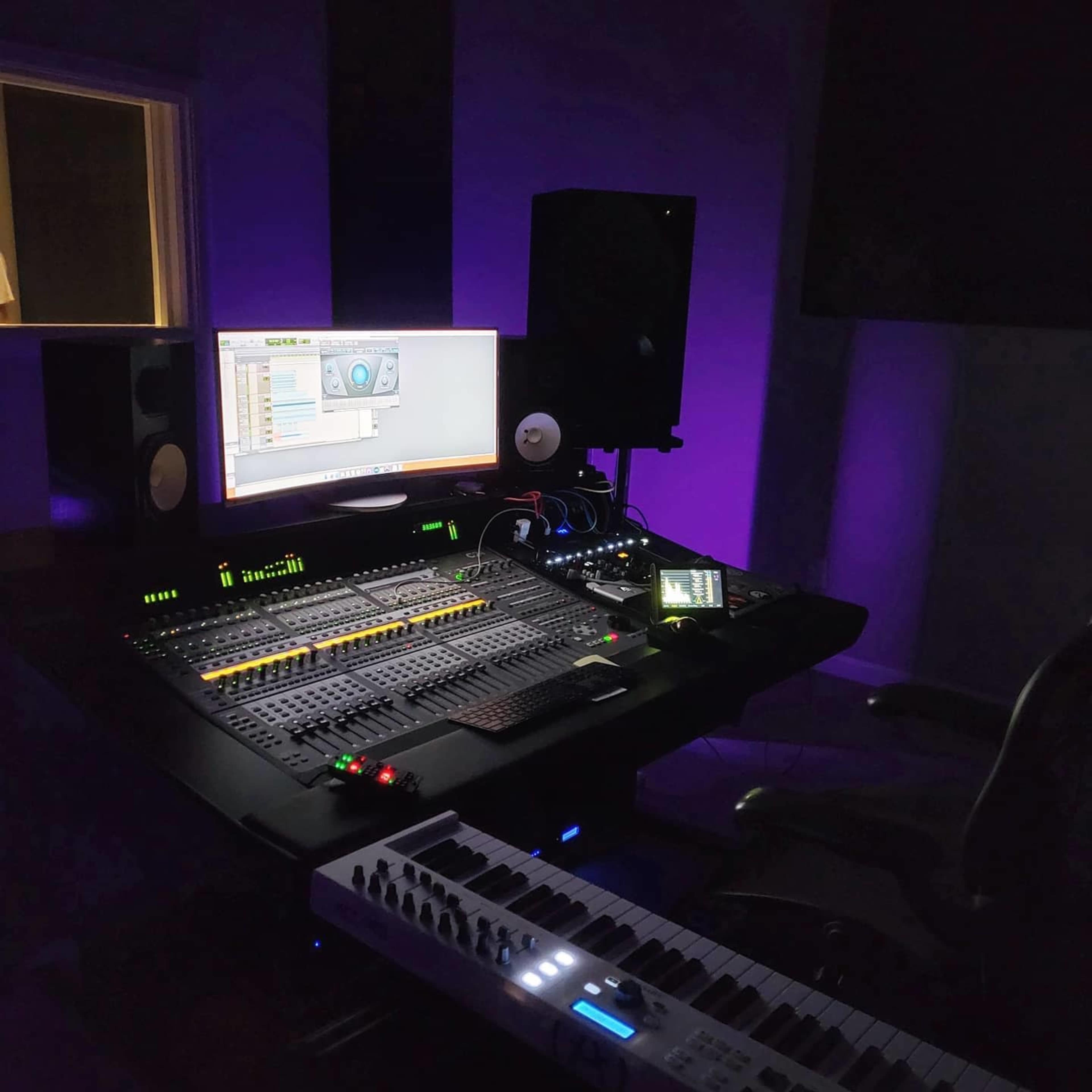 A modern music studio with a control desk, computer monitor displaying audio software, and a keyboard, all set against a dimly lit background with purple lighting.