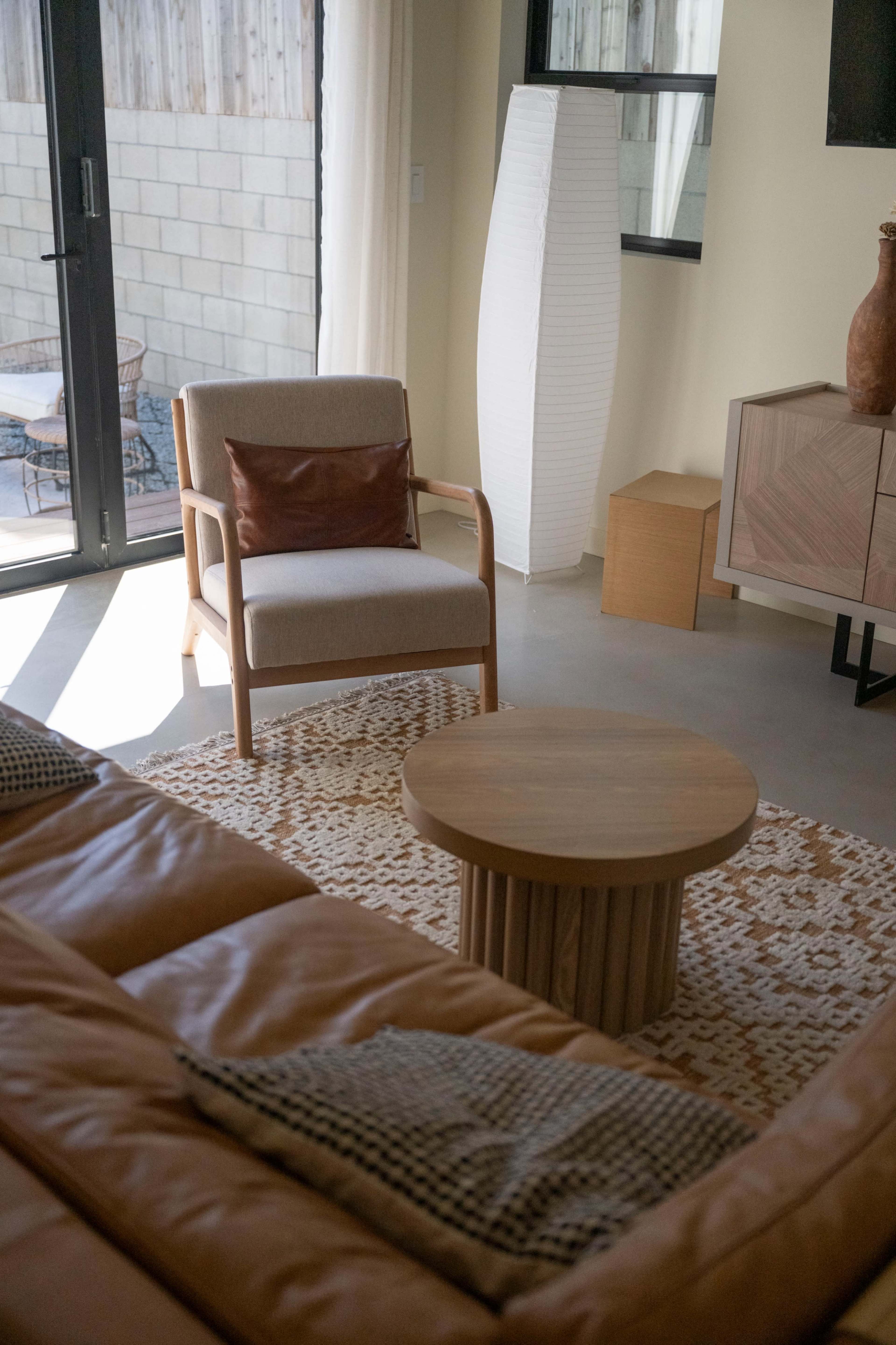 The image shows a modern living room with a brown leather sofa, a circular wooden coffee table, a wooden chair with a cushion, and a large window that provides natural light.