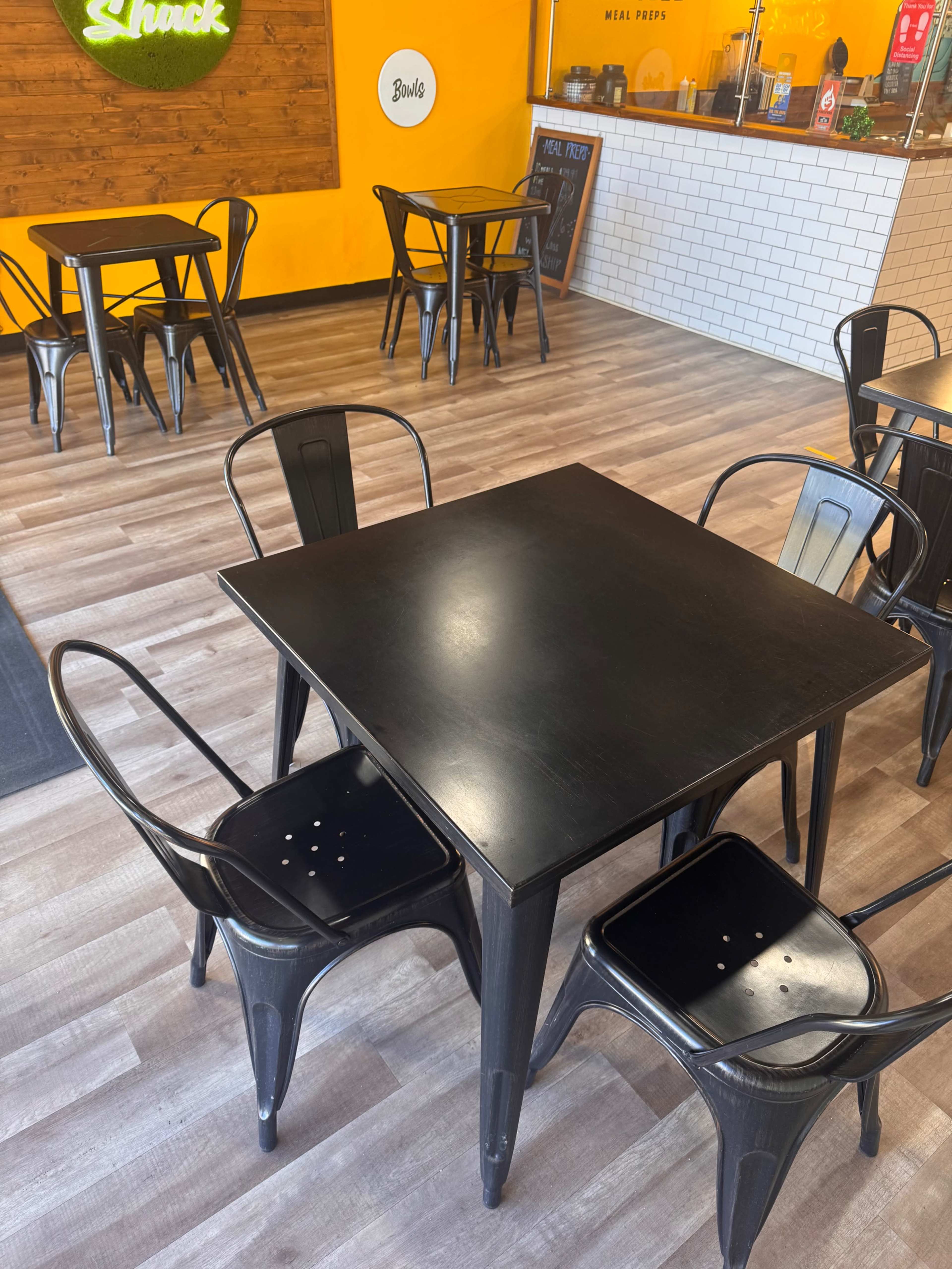 The image shows a casual dining area with black metal tables and chairs on a wooden floor, and a service counter in the background.