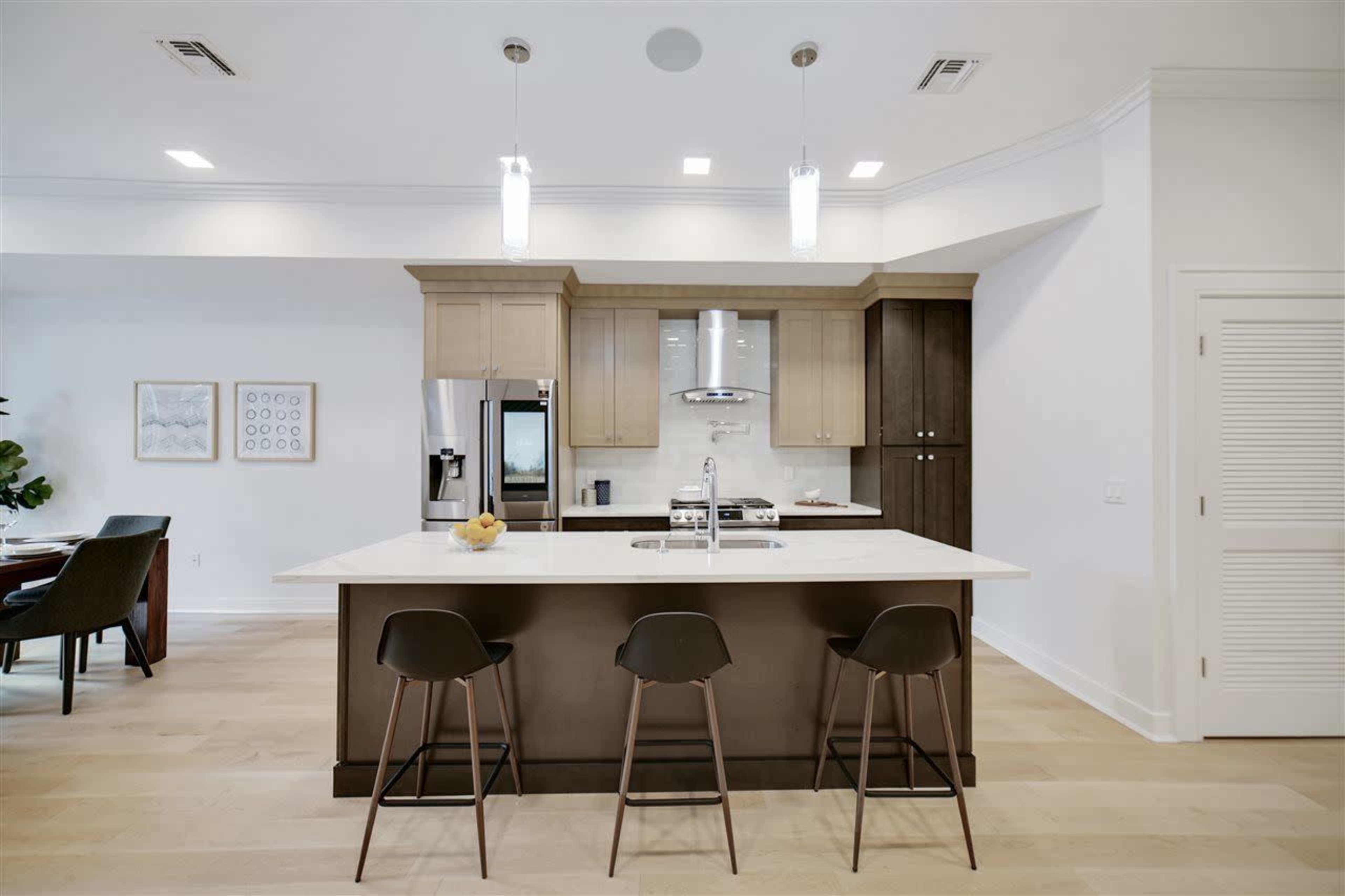 A modern kitchen features a central island with high stools, cabinetry of light and dark wood, and stainless steel appliances.