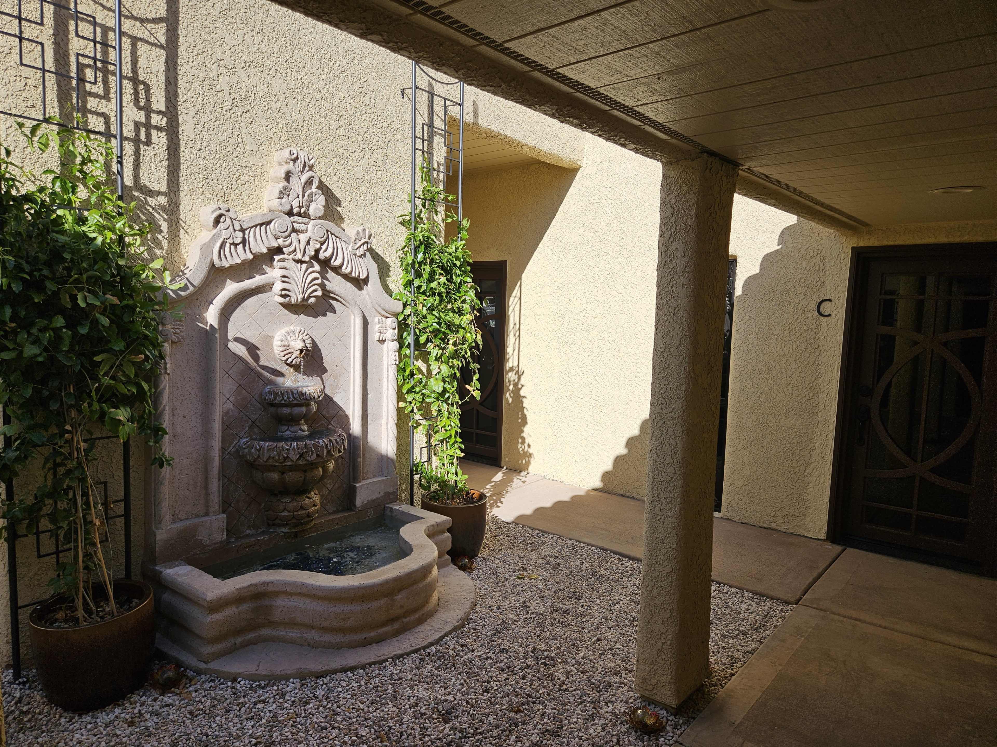 A decorative stone fountain is situated in a courtyard surrounded by potted plants, with two doors visible in the background.