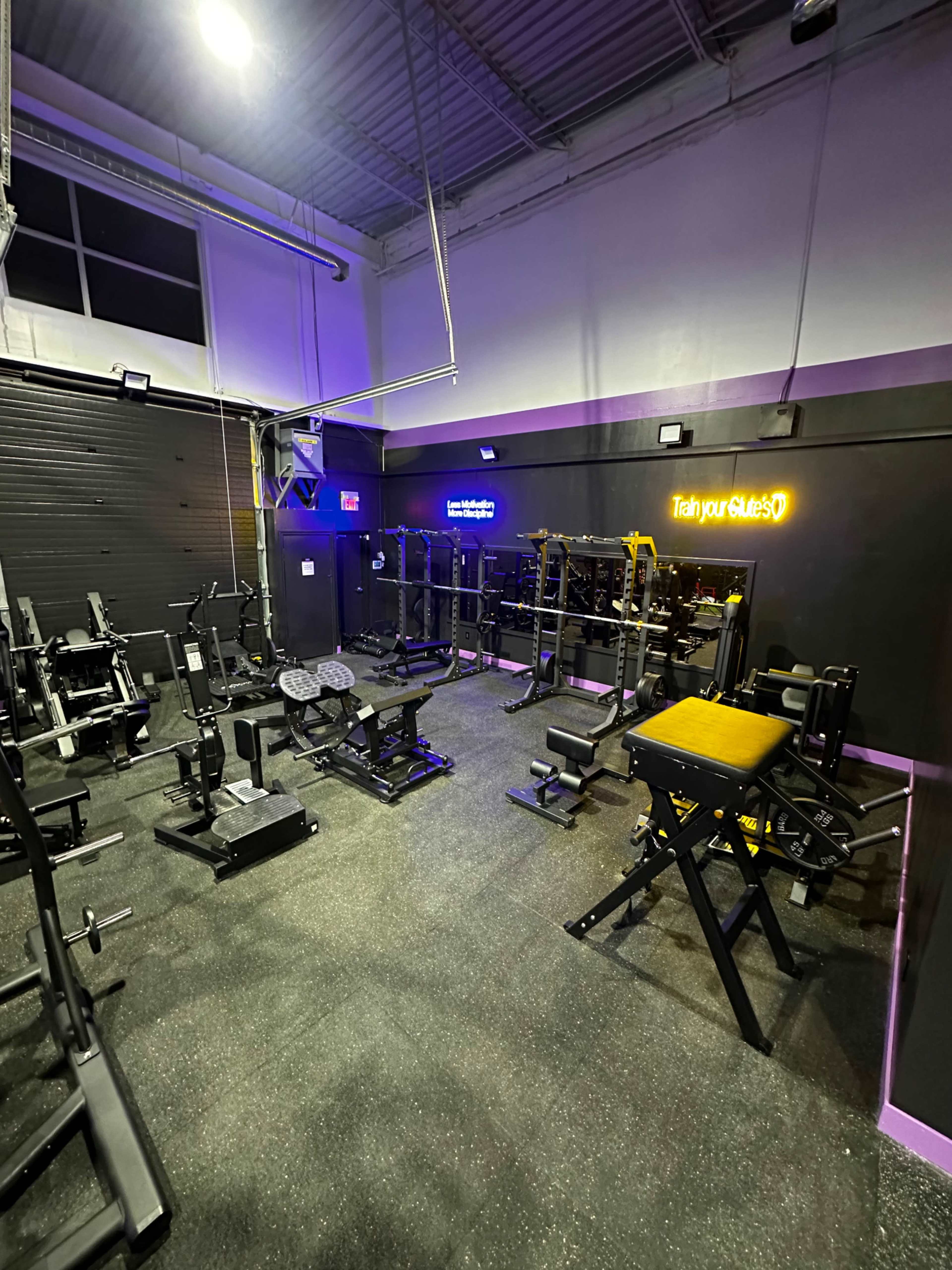 A gym equipped with various weightlifting machines and benches, illuminated by purple lighting.