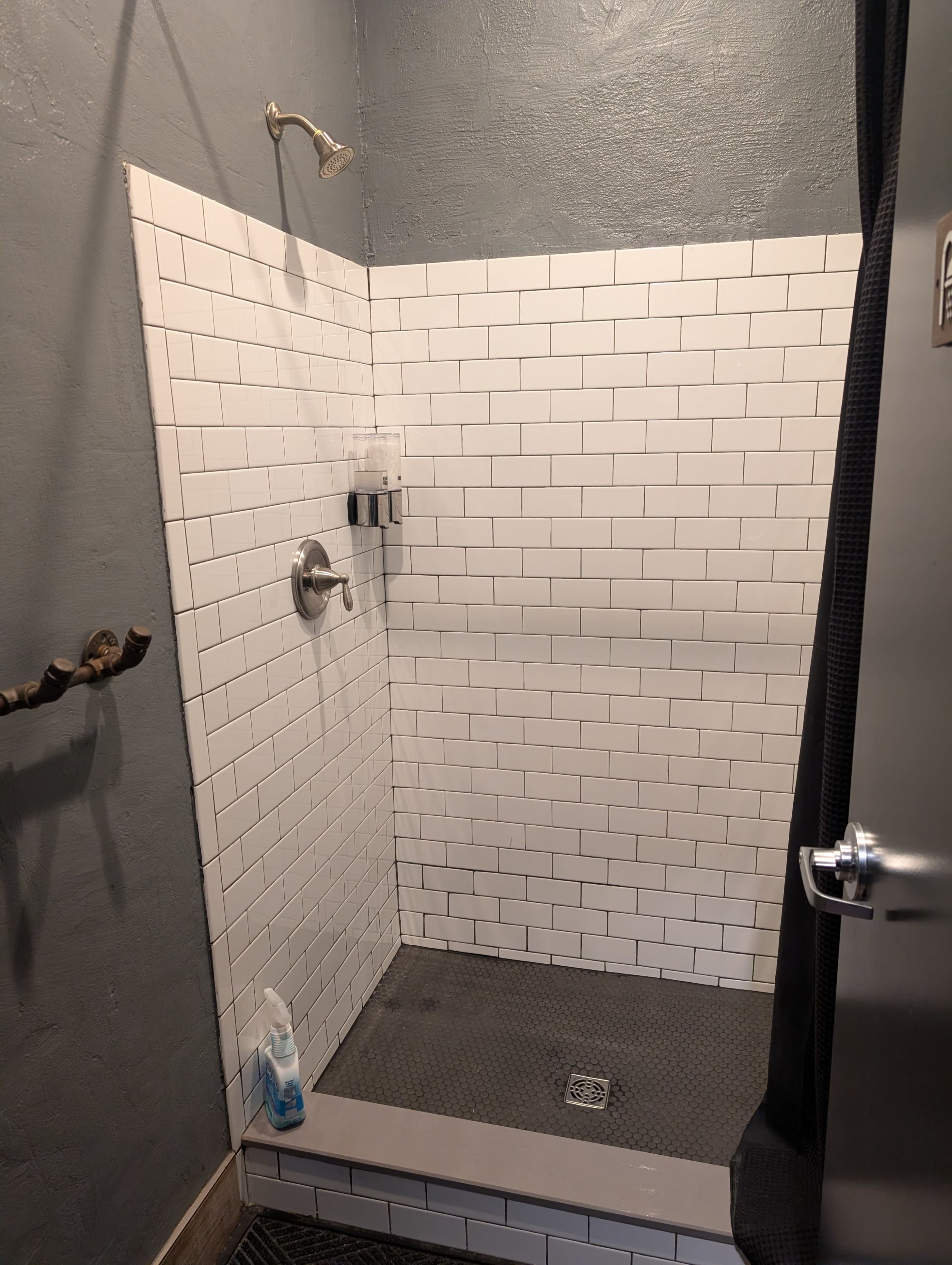 The image shows a shower area with white tiled walls, a showerhead, a soap dispenser mounted on the wall, and a black rubber mat on the floor.