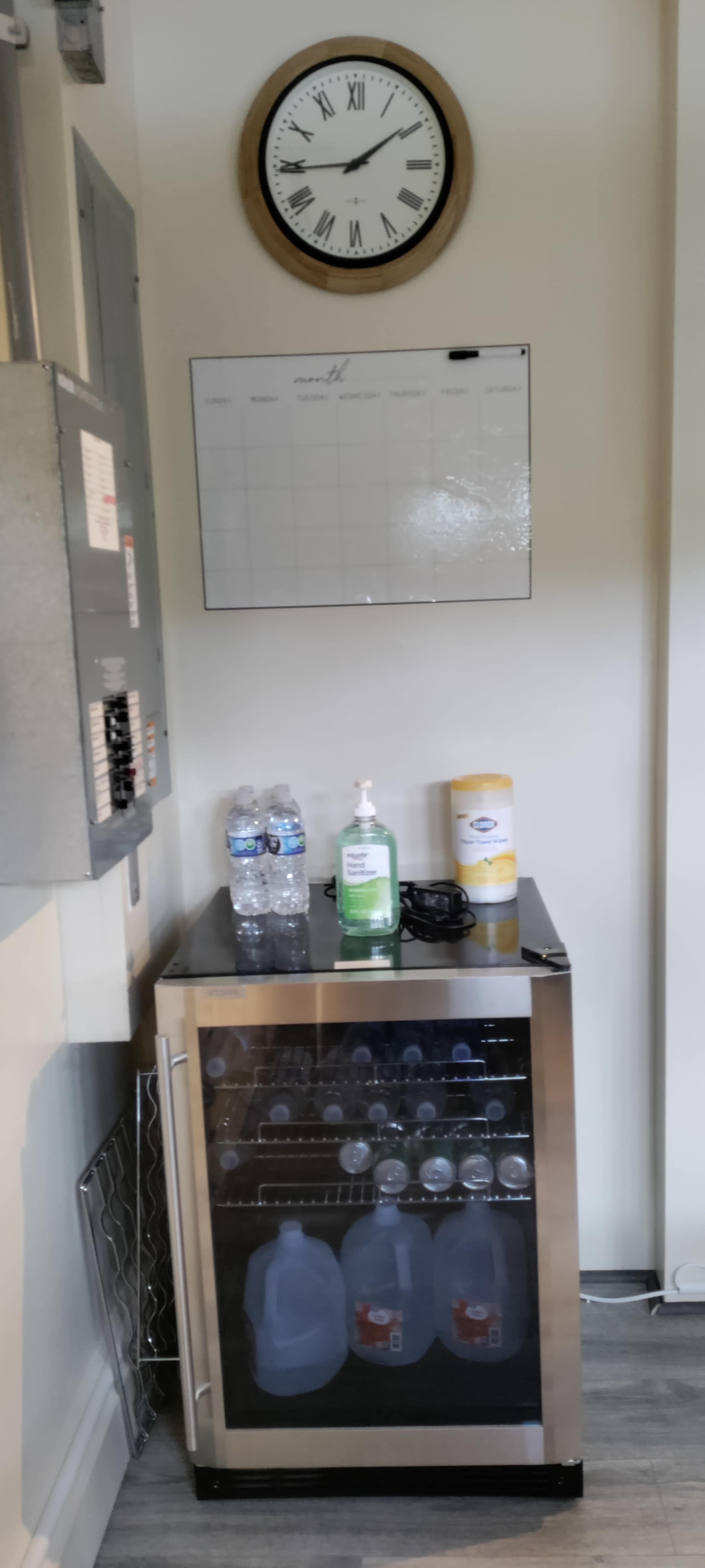A small beverage cooler filled with water bottles, hand sanitizer, and a calendar mounted on the wall above it, next to an electrical panel.