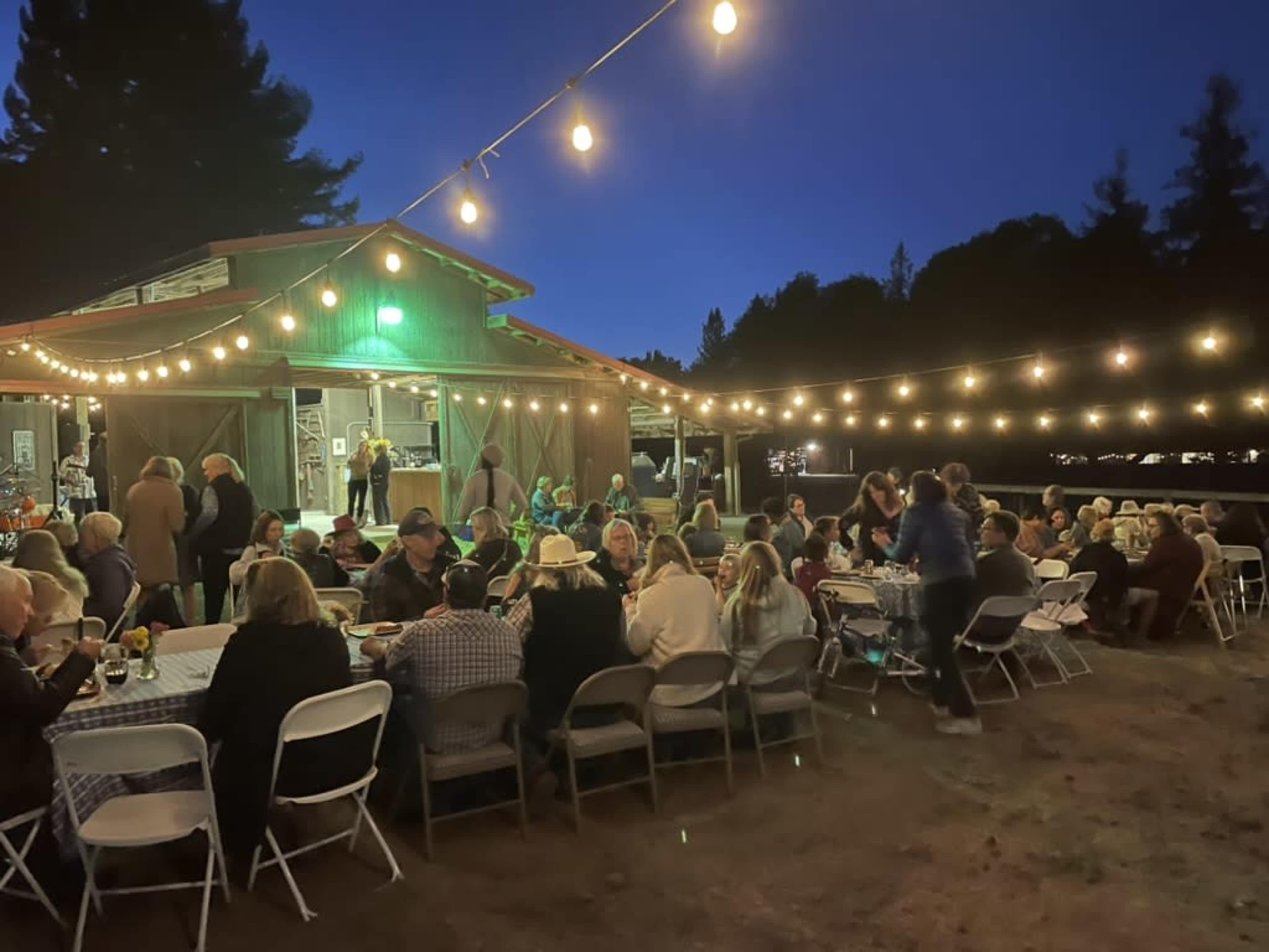 A large group of people is gathered at tables for an outdoor dinner party in front of a rustic building illuminated by string lights.