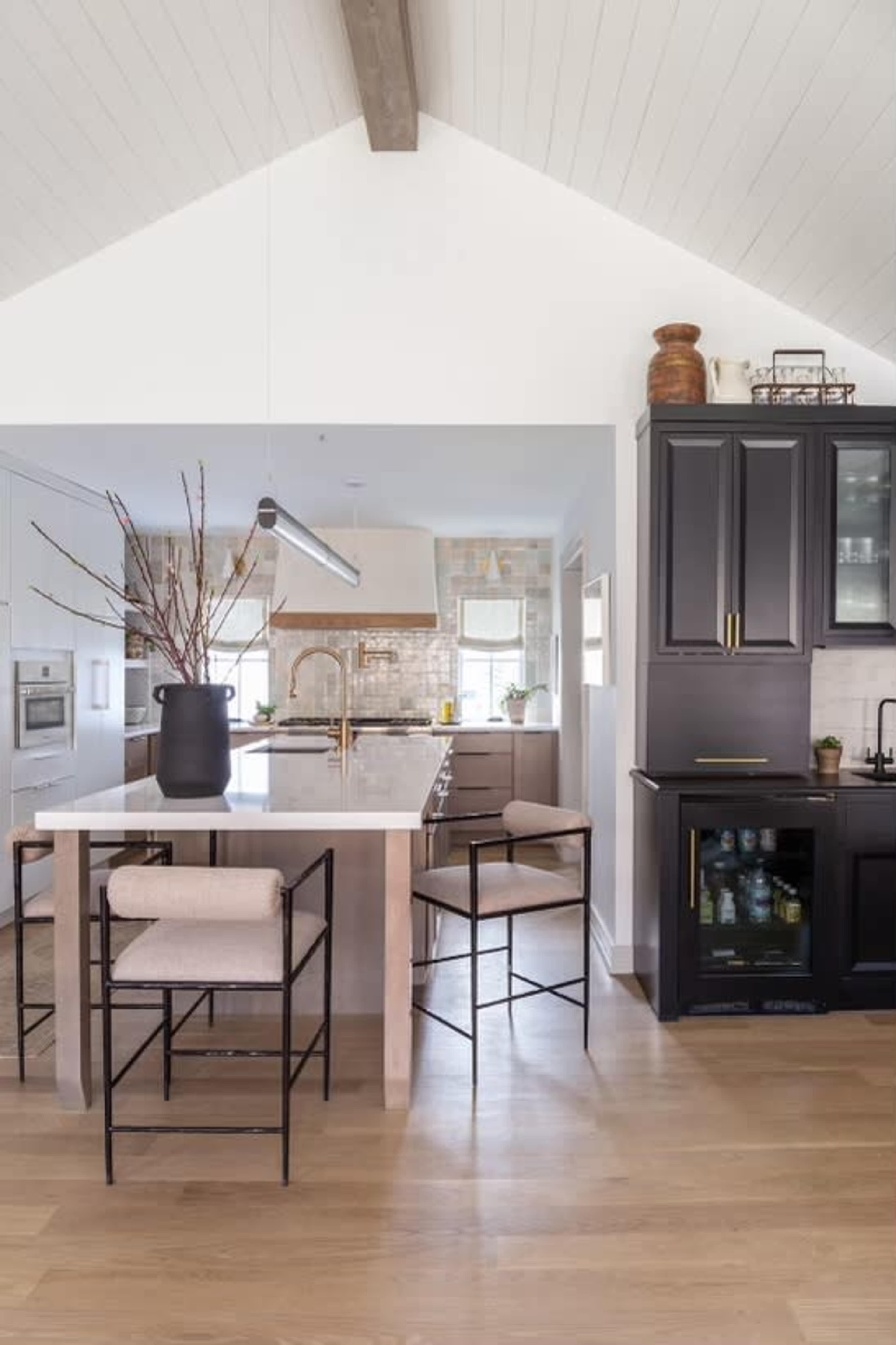 A modern kitchen features a large island with bar stools, a cabinetry section with glass doors, and a high ceiling with exposed beams.