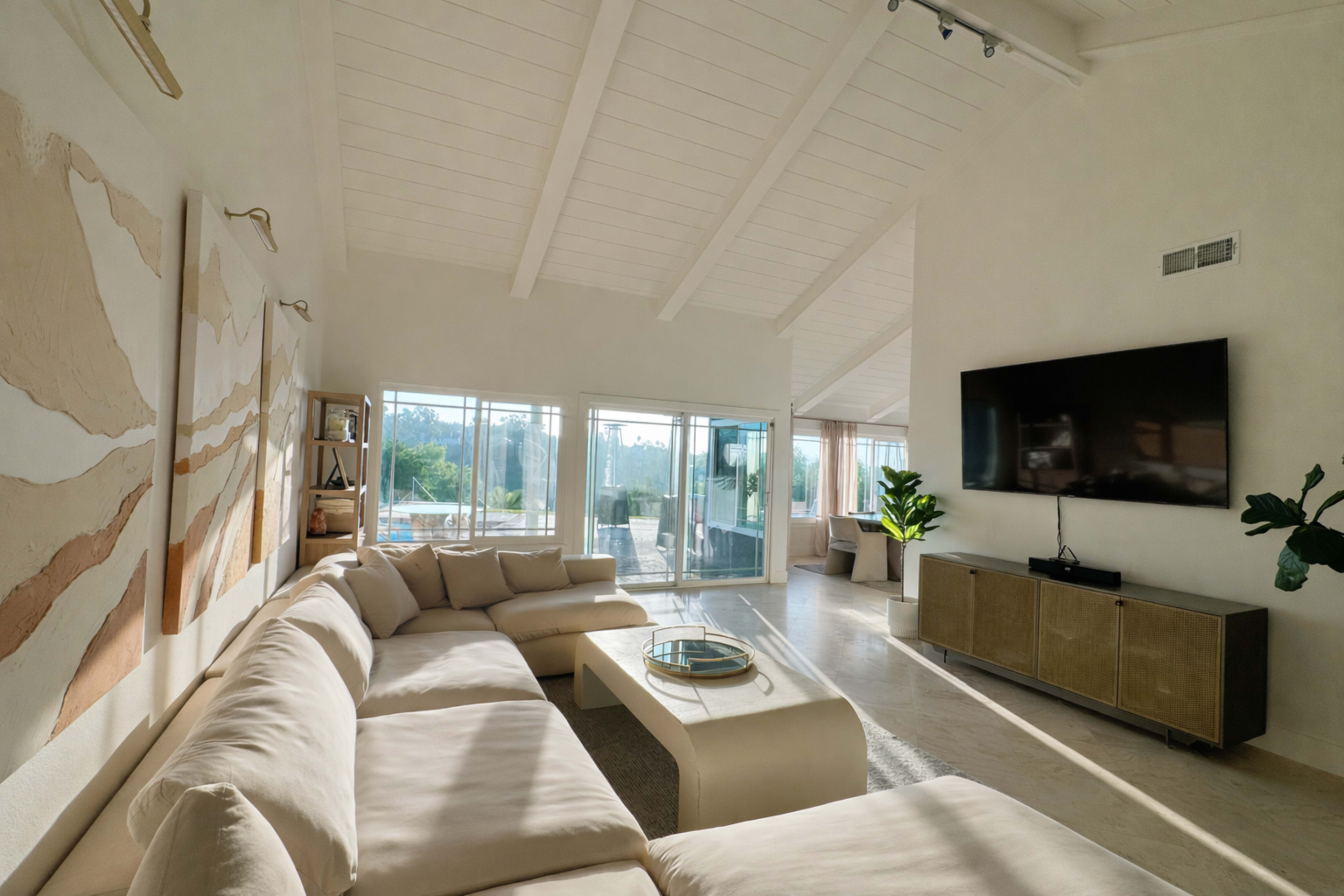 The image shows a modern living room with a large sectional sofa, a coffee table, a TV mounted on the wall, and large windows that allow natural light to fill the space.