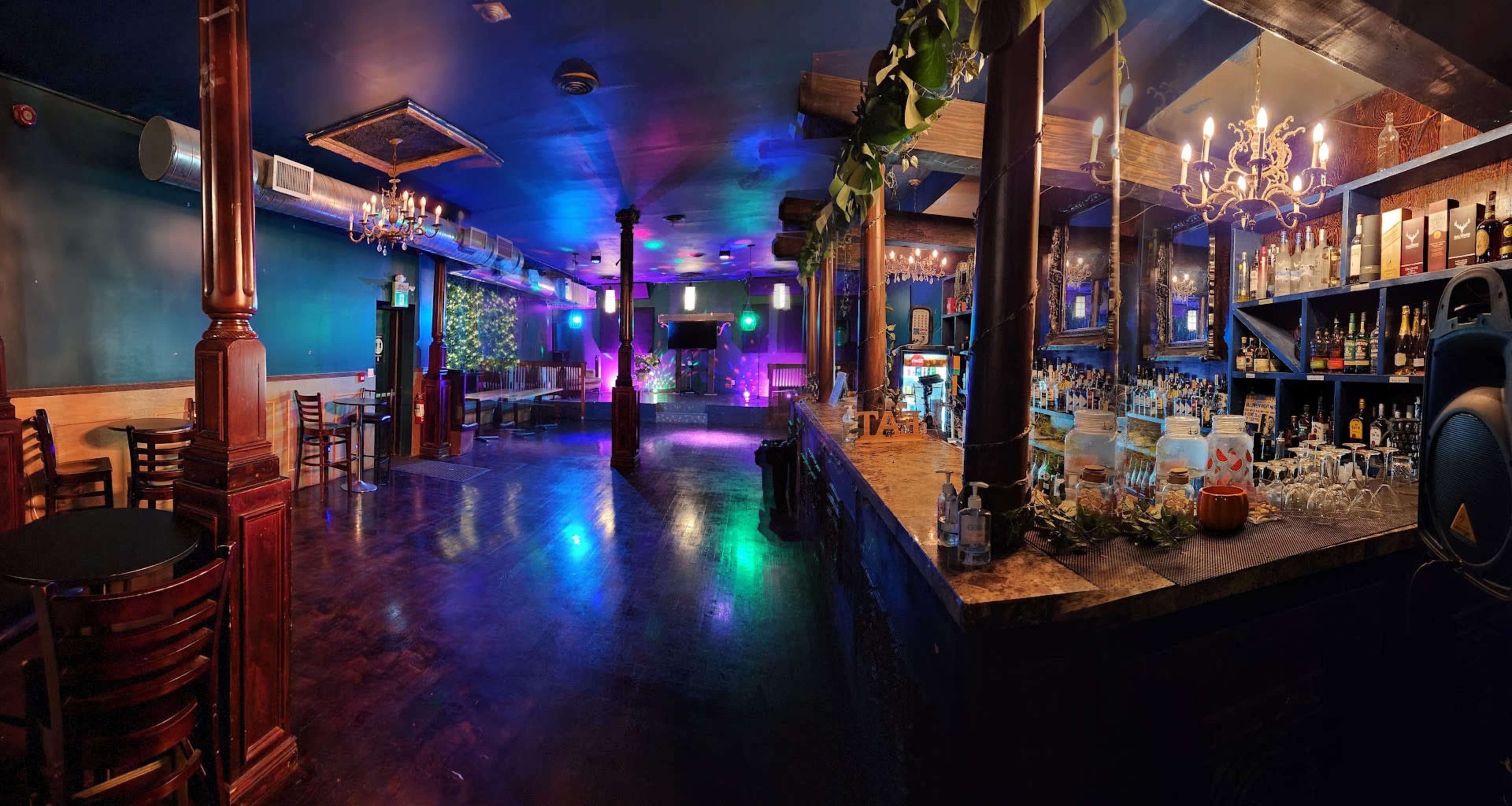 A spacious bar area with a wooden counter, various bottles displayed on shelves, and colorful lights illuminating the room.