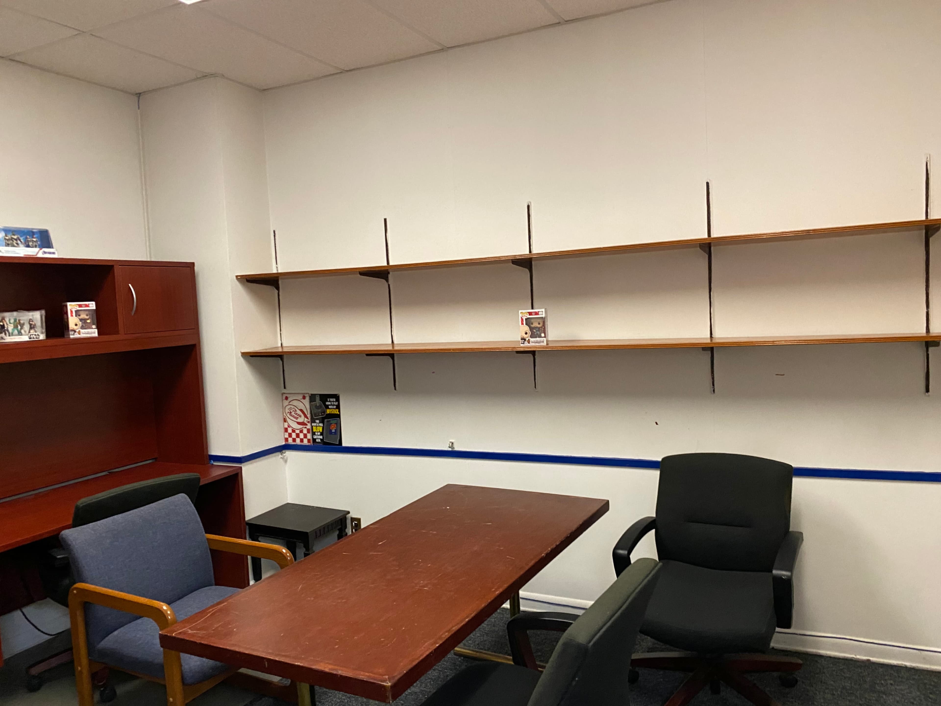 The image shows a small office space with a wooden table, three chairs, a shelving unit on the wall, and a red storage cabinet.