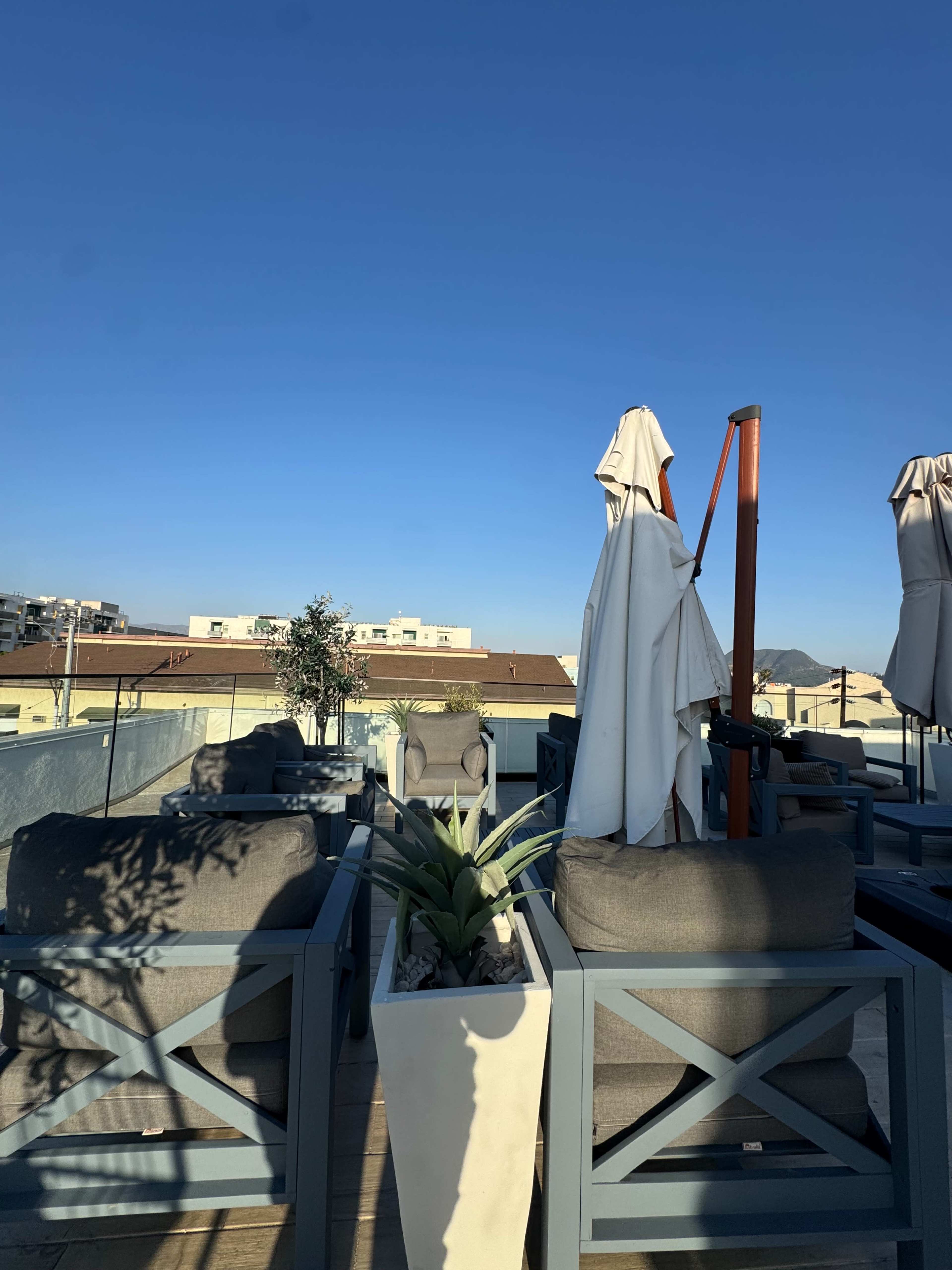 A rooftop seating area features gray sofas, potted plants, and several umbrellas against a clear blue sky.