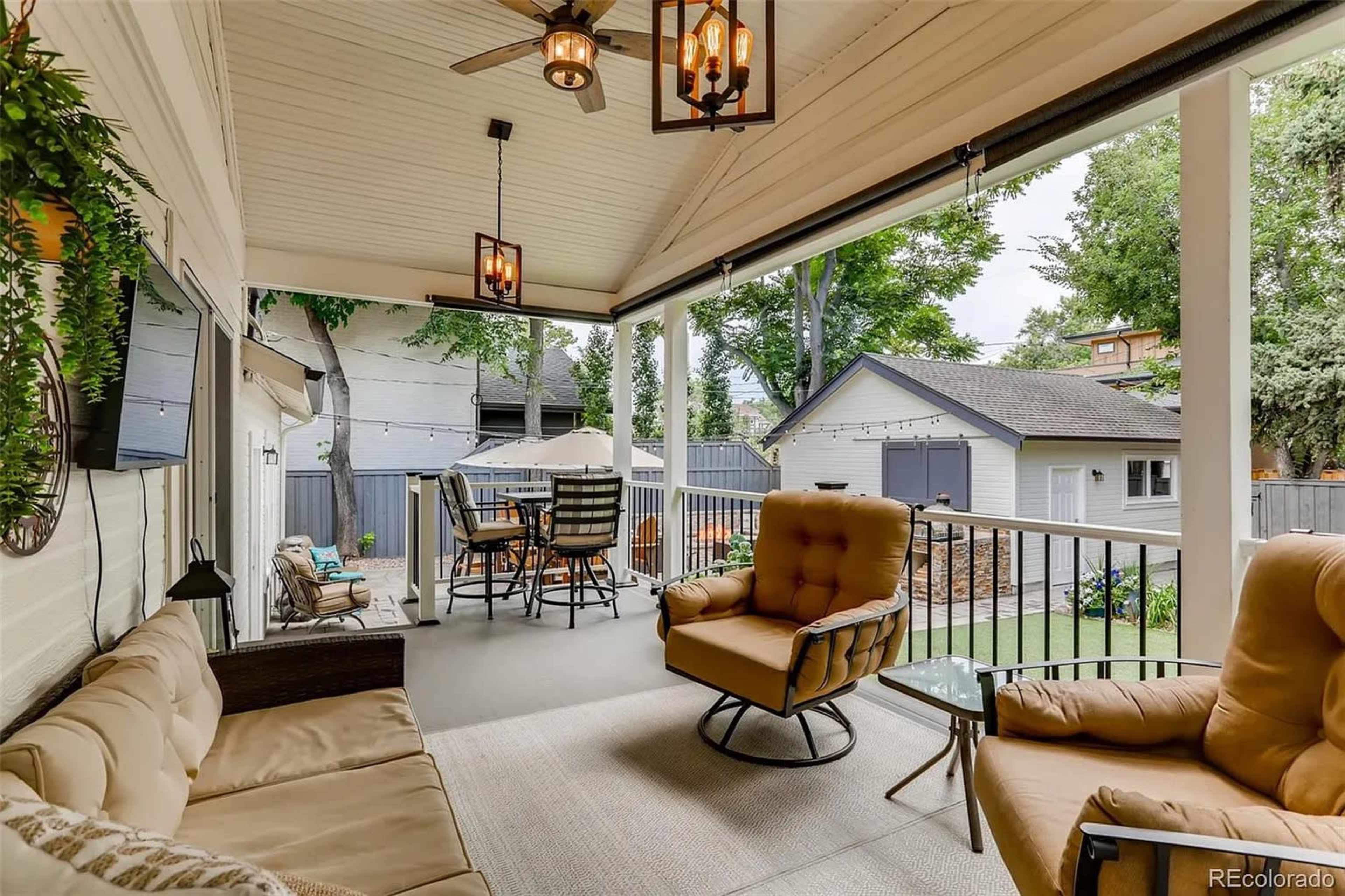 The image shows a covered patio with comfortable seating, a television mounted on the wall, and a dining area that overlooks a backyard with a shed.
