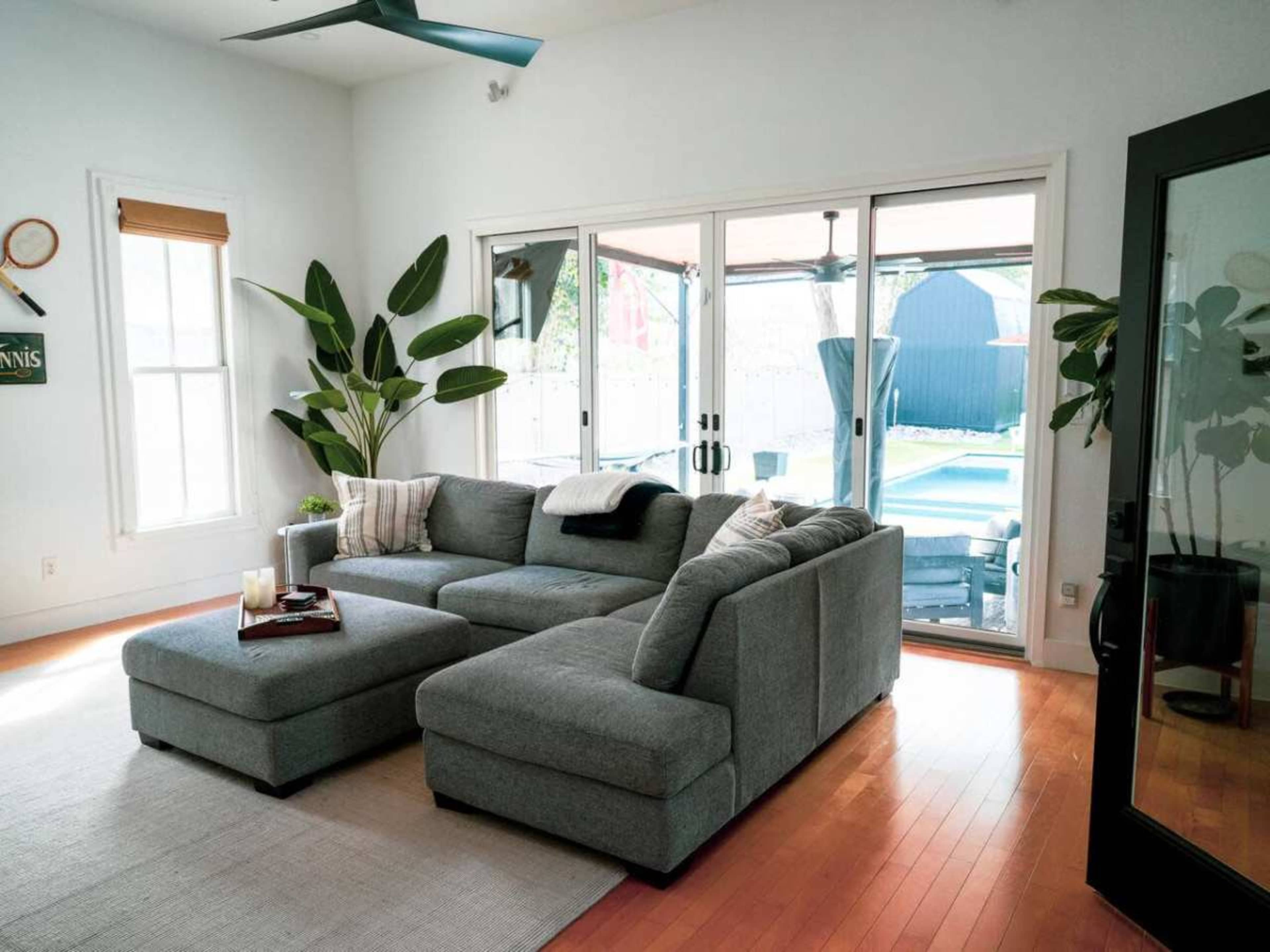 The image shows a spacious living room featuring a large gray sectional sofa, a coffee table with candles, and glass doors leading to an outdoor area with a pool.