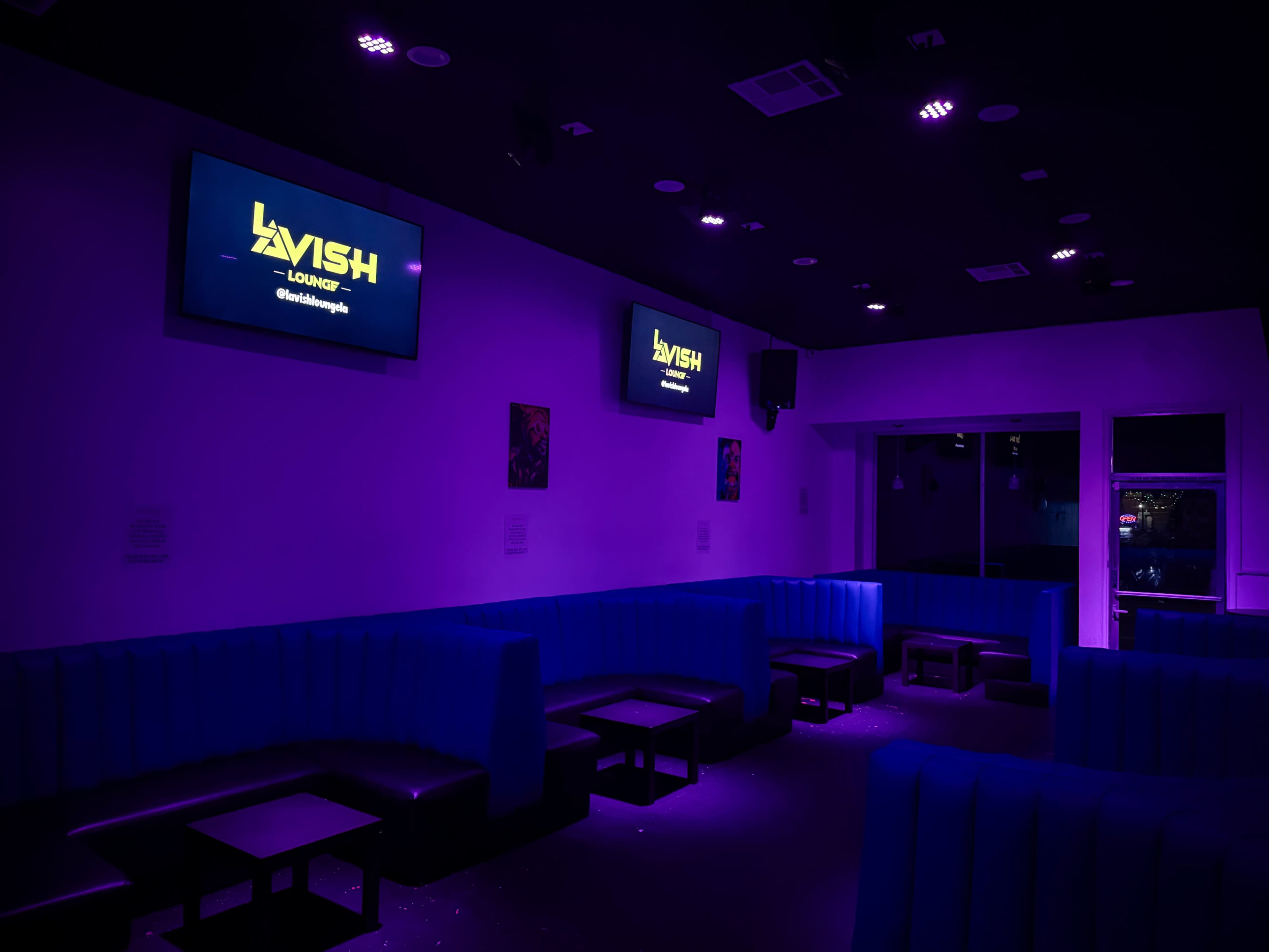 The image shows a nightclub interior with purple lighting, featuring blue seating areas and two screens displaying the word "LAVISH" on the walls.