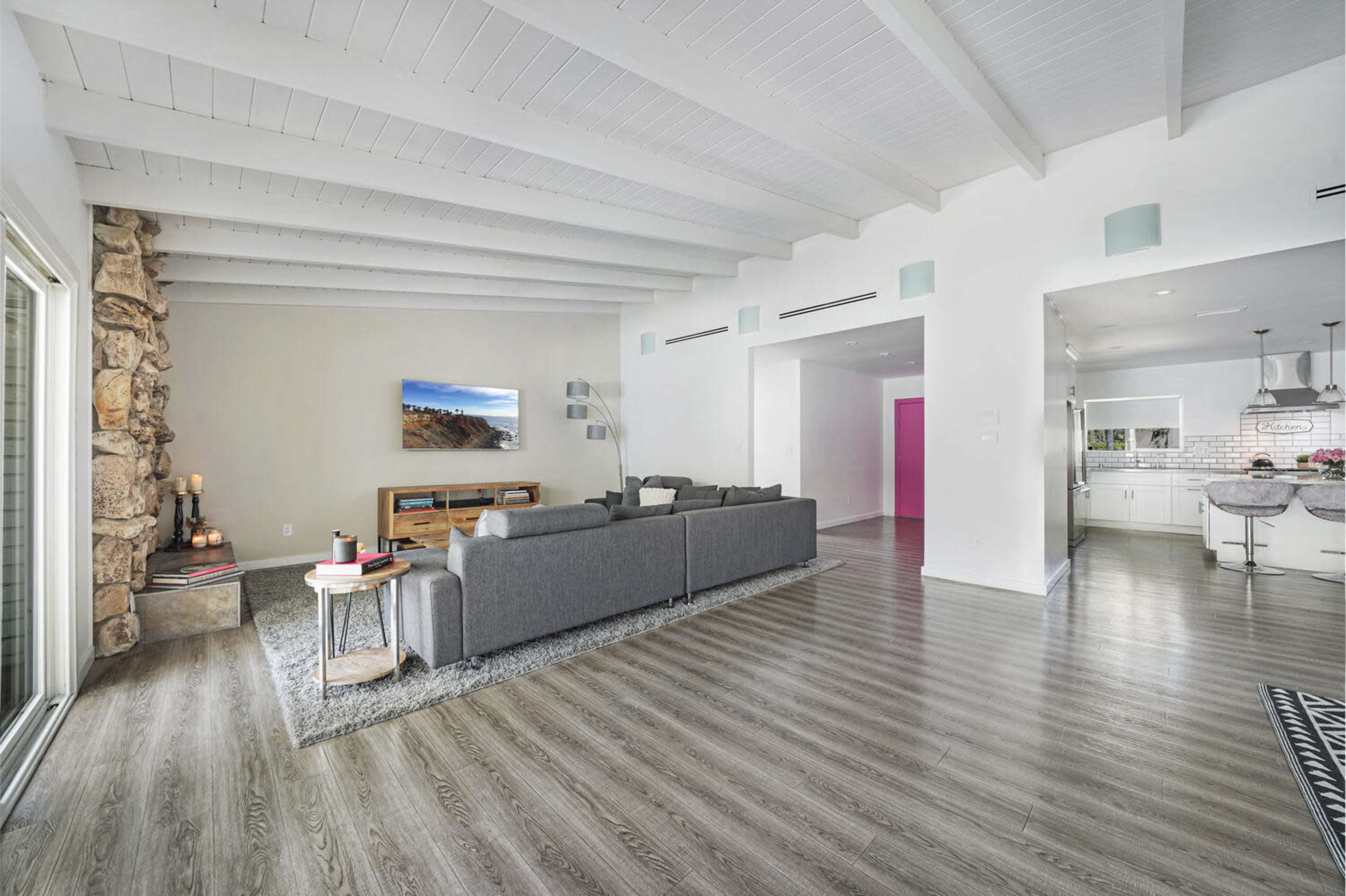 A spacious living area features a gray sectional sofa, wooden media console, and open sightlines toward a modern kitchen with white cabinetry.