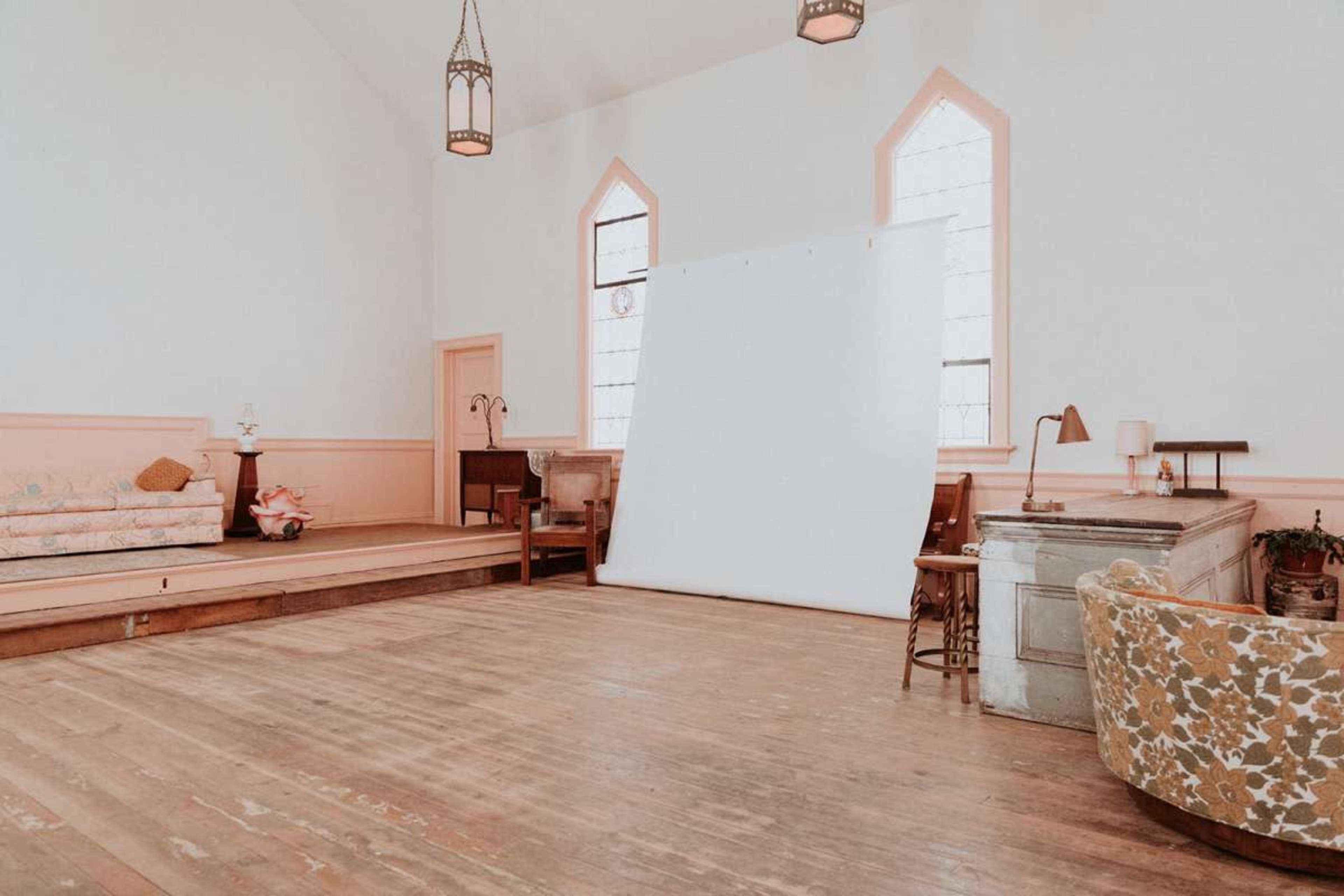 The image shows a spacious, well-lit room with wooden floors, featuring a large white backdrop and various seating arrangements, including a sofa and chairs.