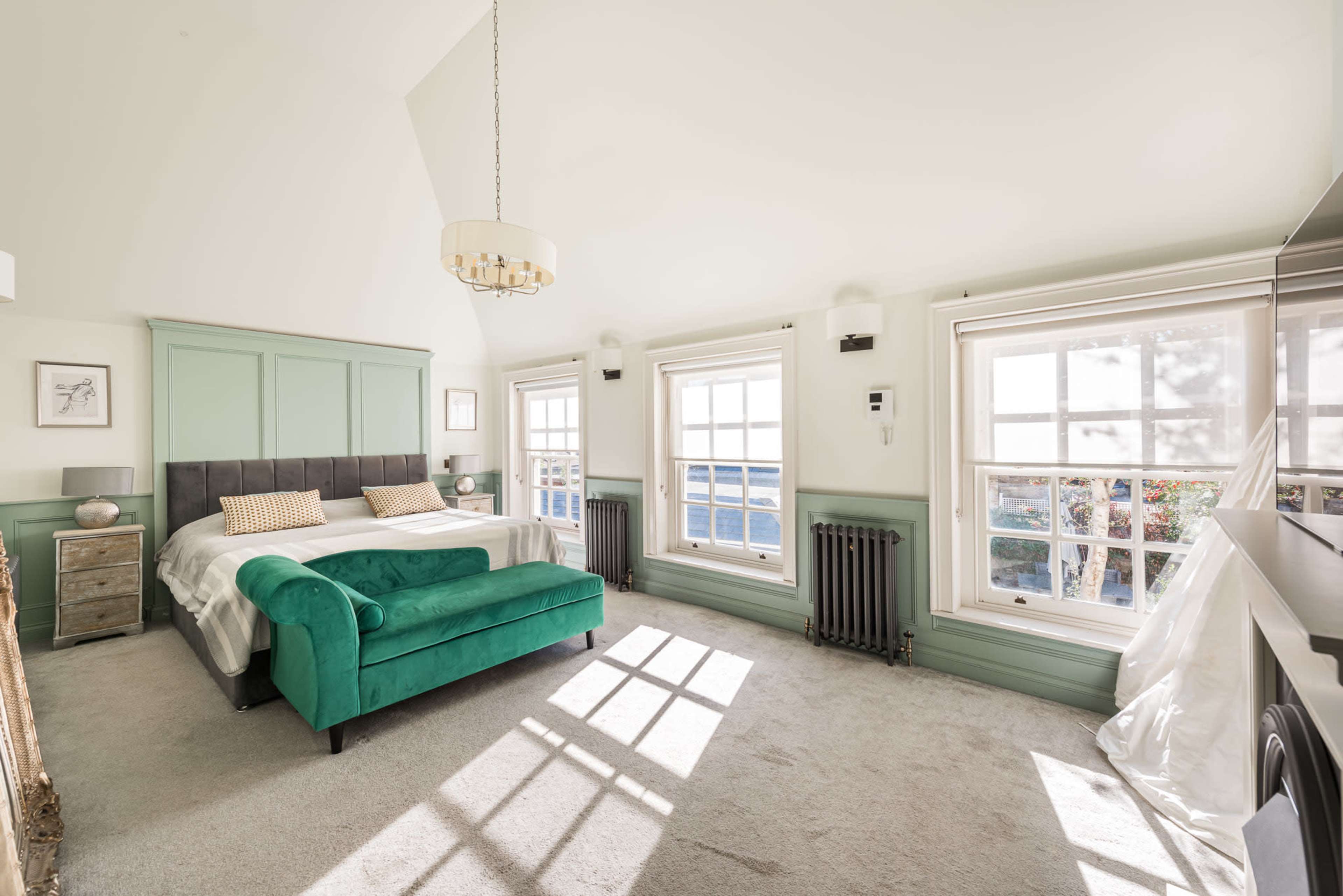 The bright bedroom features a large bed with green accents, two windows with natural light, and a plush carpeted floor.