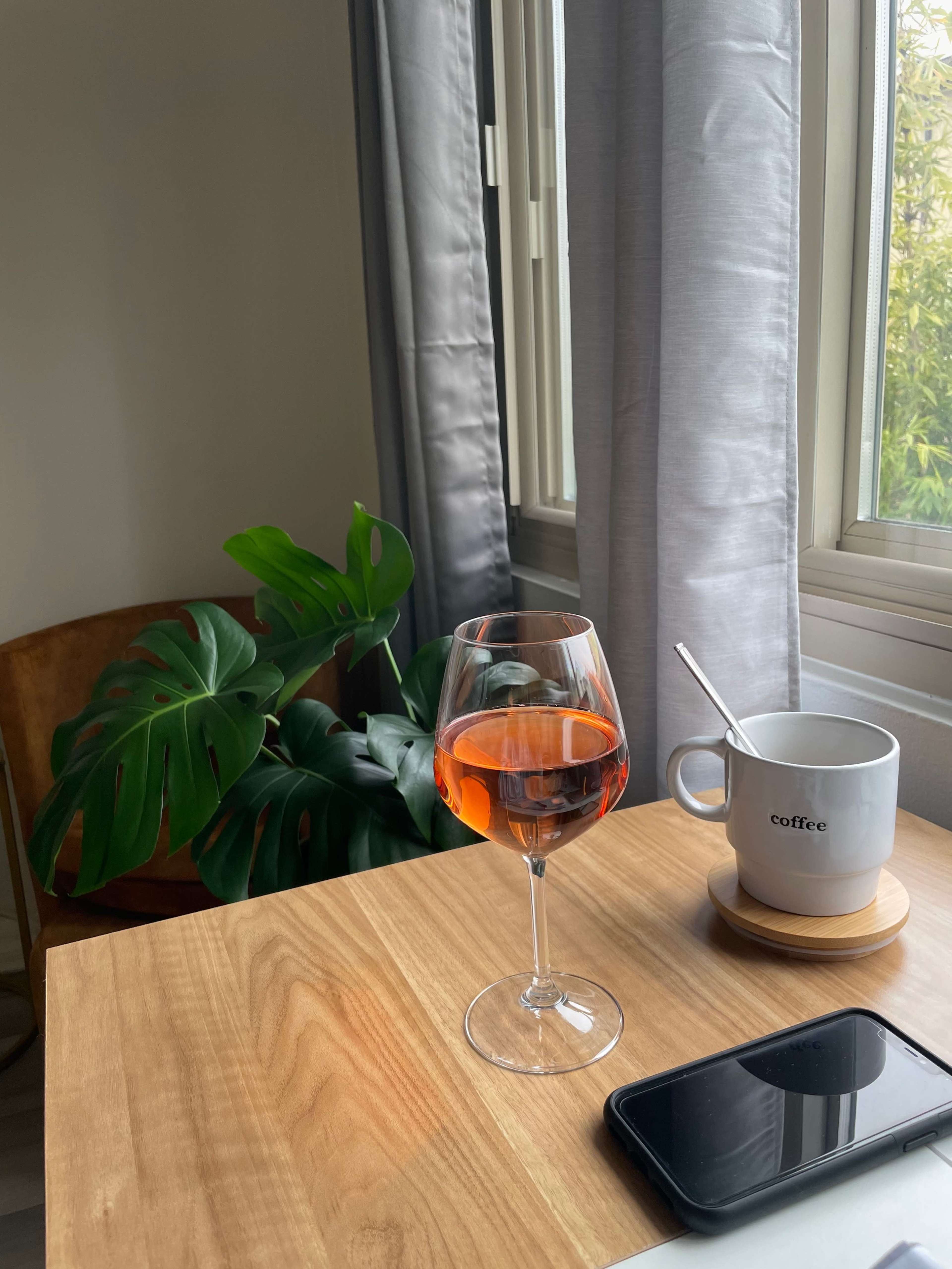 A glass of rosé wine sits on a wooden table next to a coffee cup and a smartphone, with a large green plant in the background.