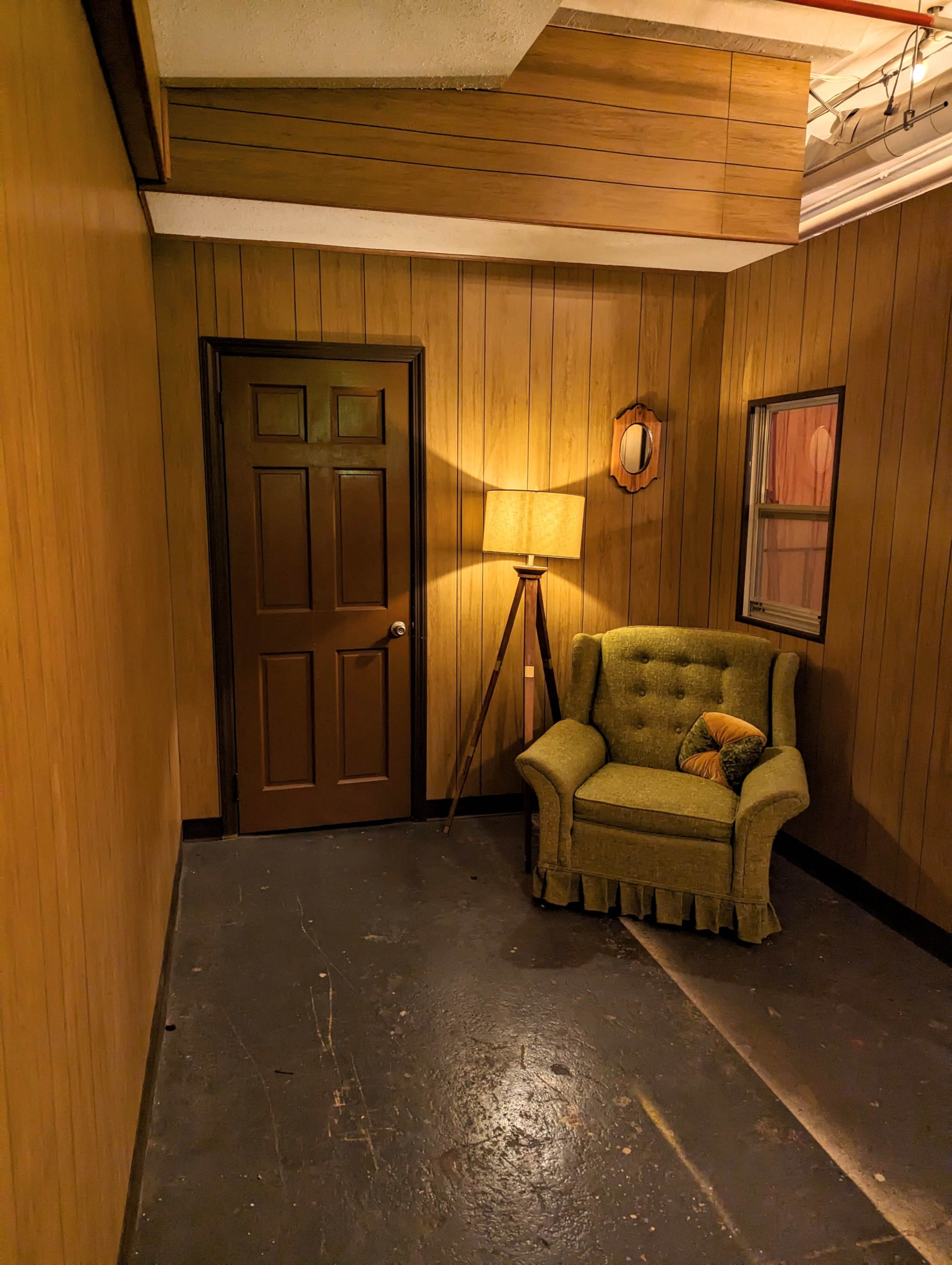 A small, dimly lit room with wood-paneled walls, a green upholstered chair, a floor lamp, a mirror, and a closed door.