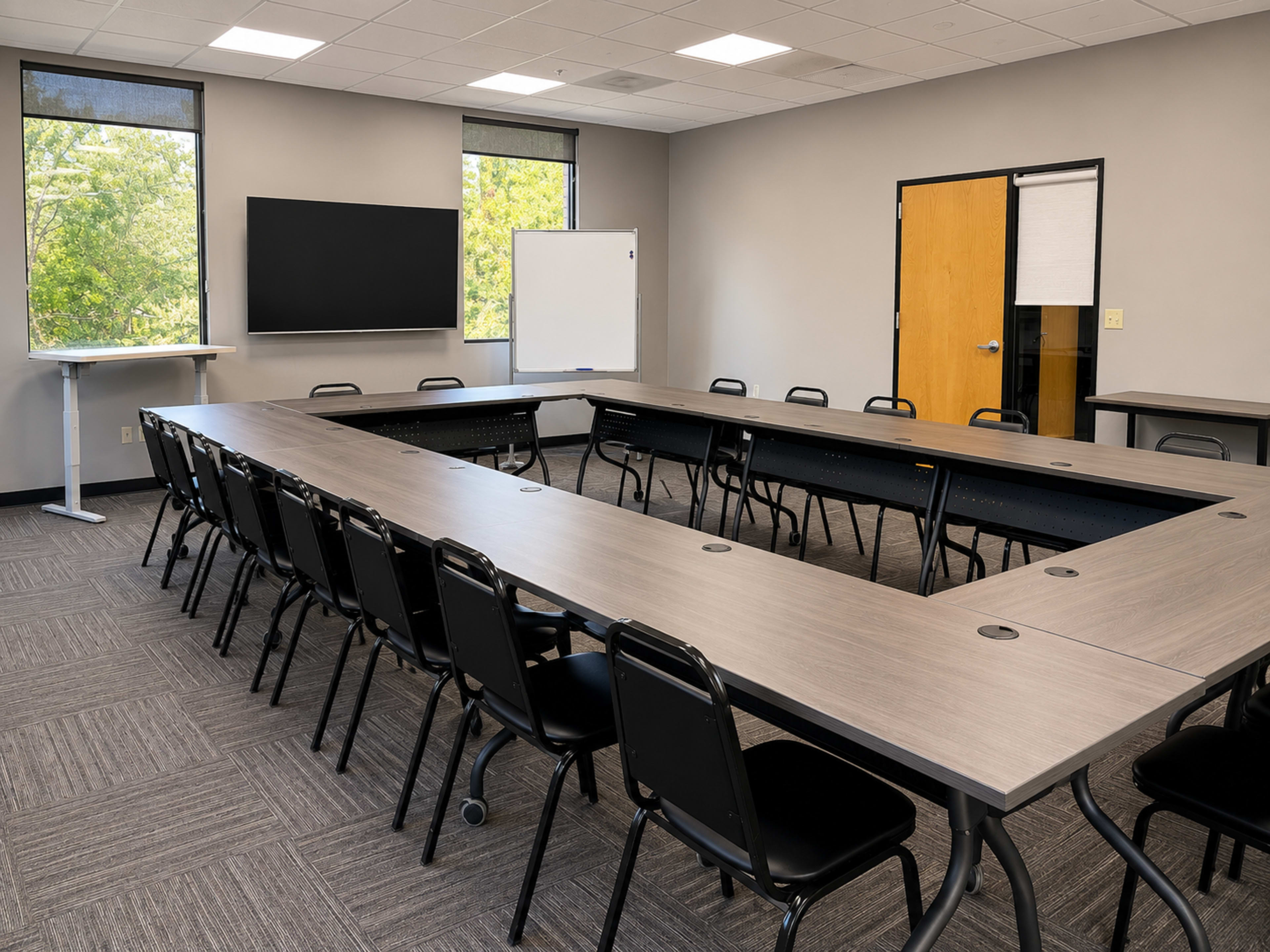 A rectangular conference room features a large table surrounded by black chairs, with windows providing natural light and a whiteboard and screen at one end.