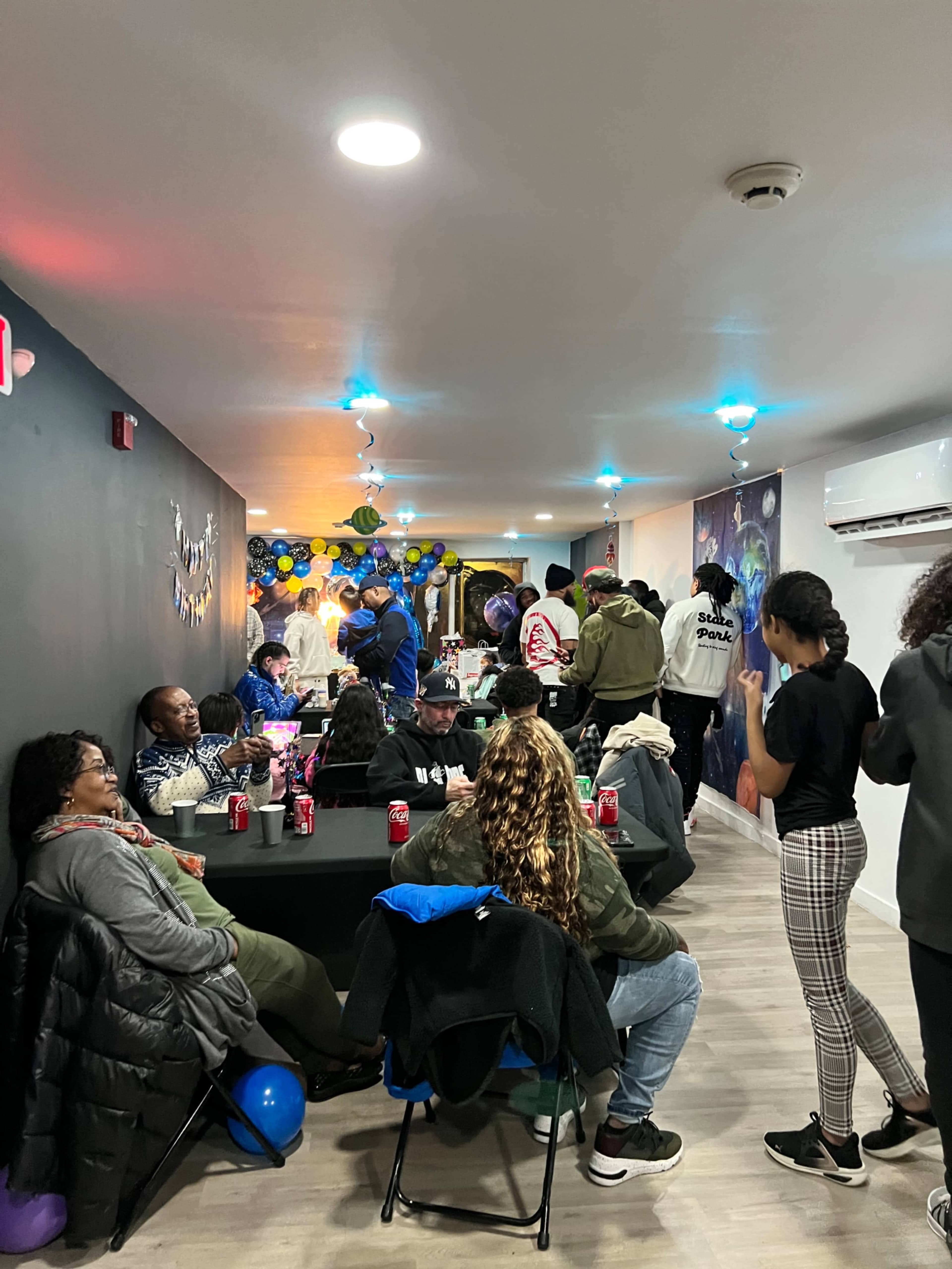 A group of people gathers in a decorated room with tables and colorful balloons for an event, while some are seated and others are mingling.