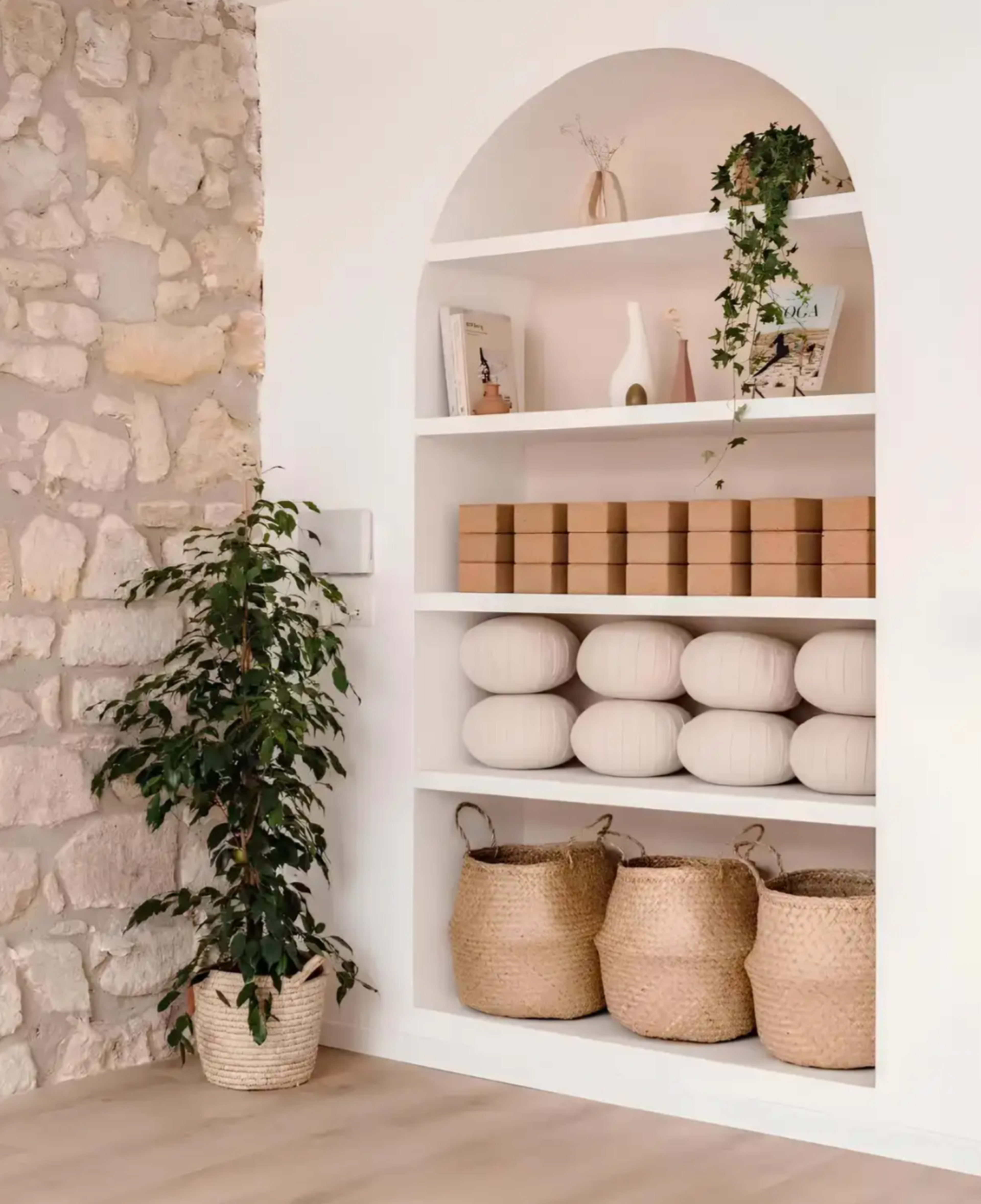 The image shows a minimalist shelf built into a wall, displaying decorative items, stacked boxes, and cylindrical cushions, accompanied by a potted plant and woven baskets at the bottom.