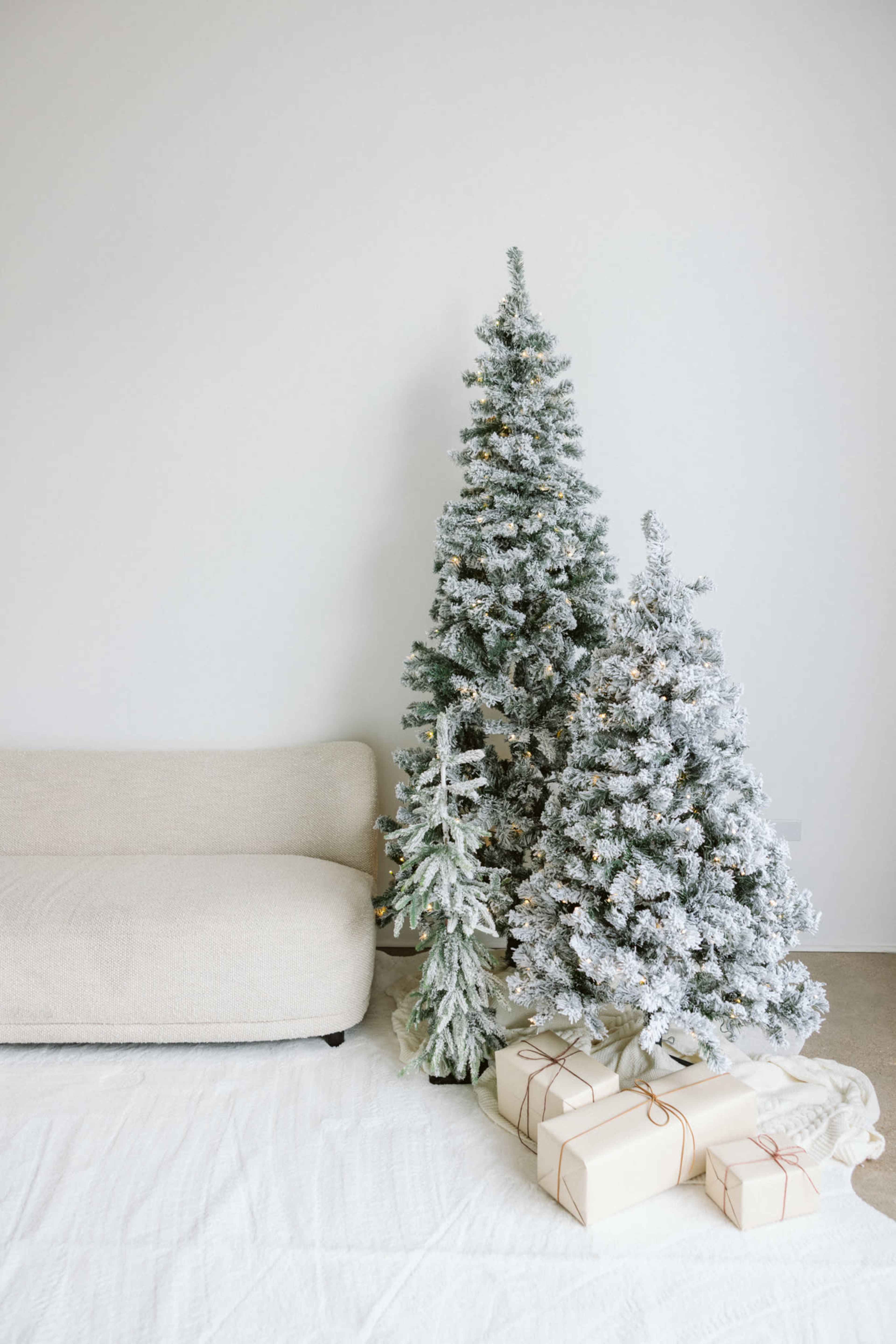 A cozy living room features two snowy Christmas trees beside a wrapped stack of gifts and a beige sofa.