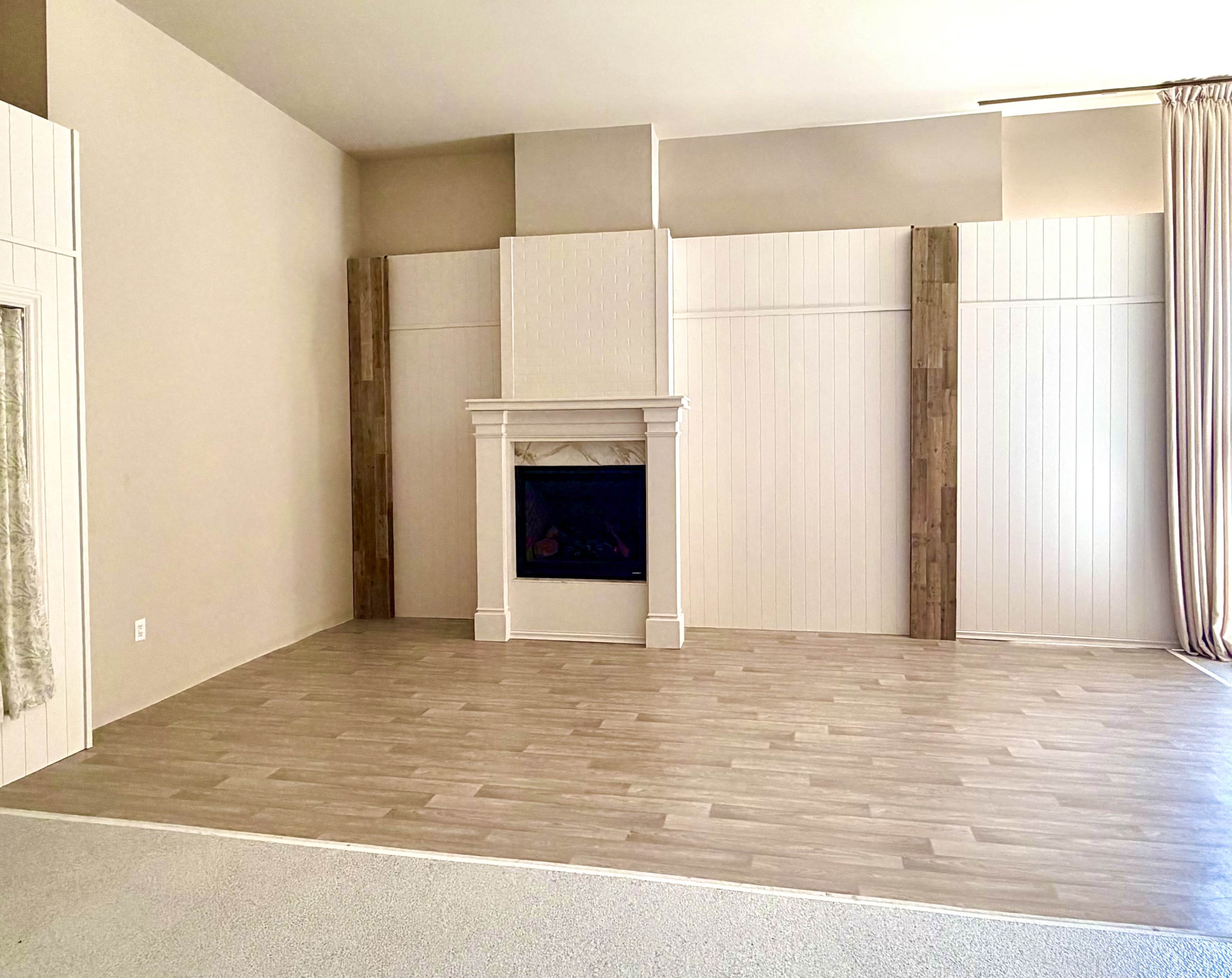 The image shows a spacious, undecorated living room with a fireplace centered on a light-colored wall and wood accents on either side.