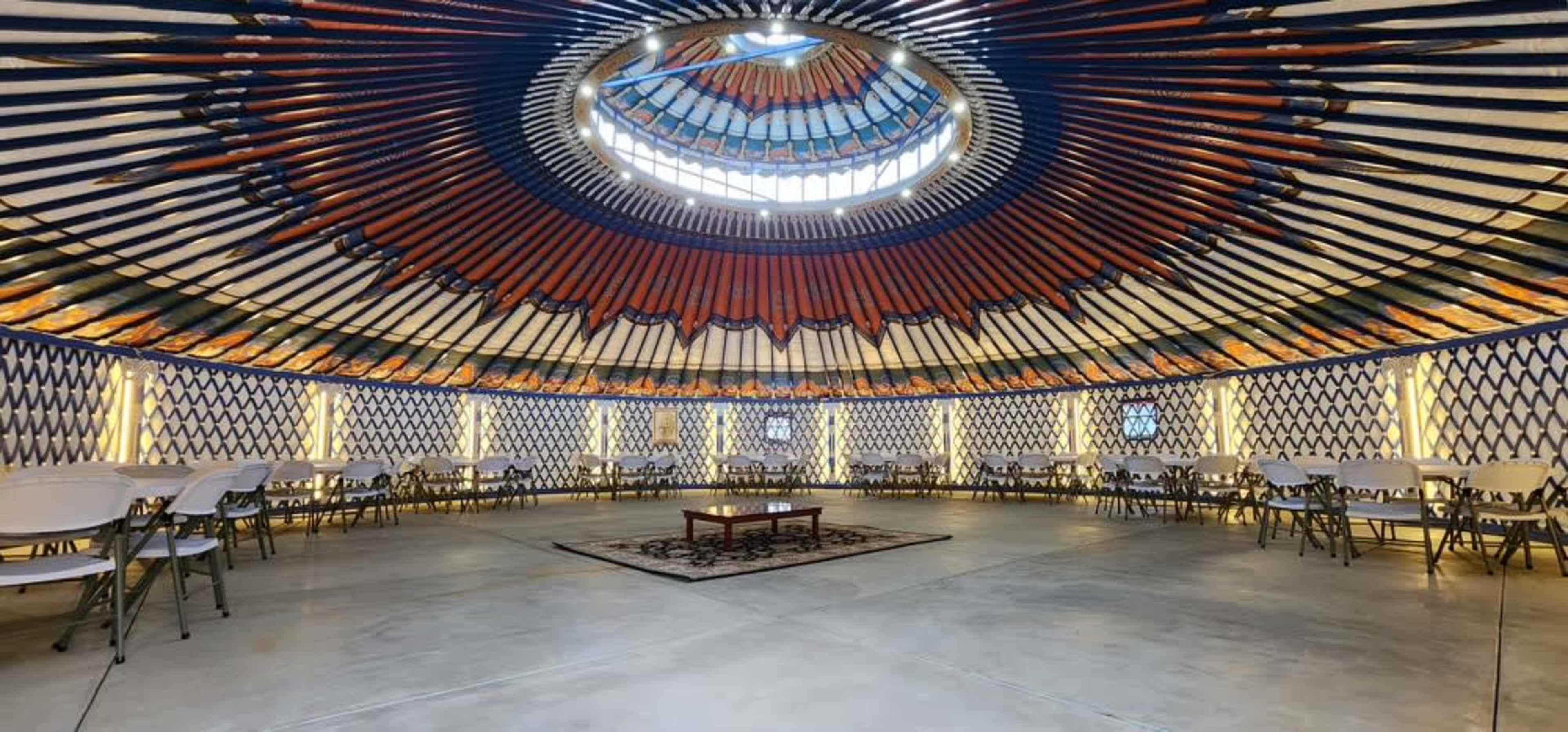 The image shows the interior of a yurt with a circular ceiling and rows of folding chairs arranged around a central table.