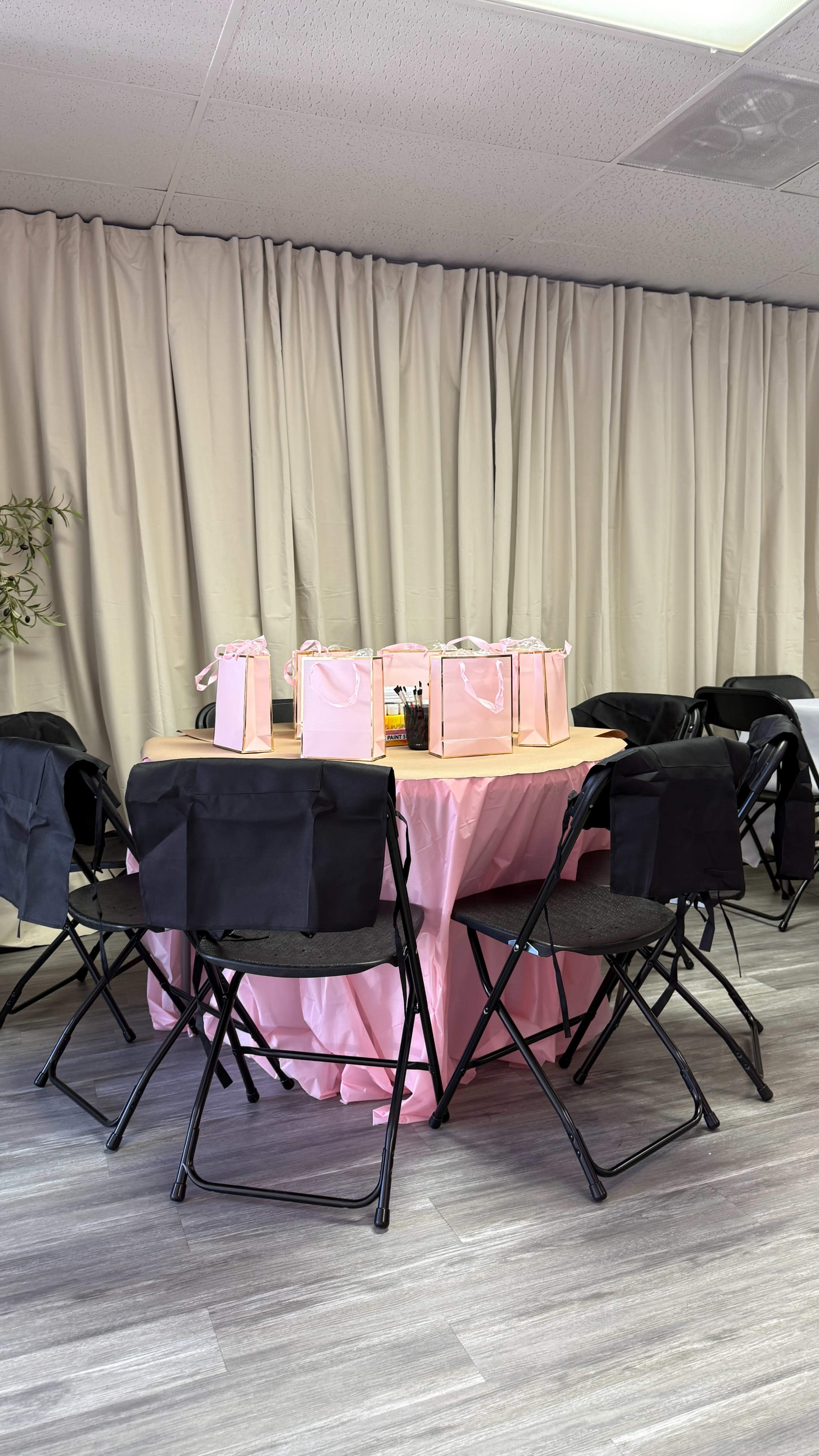 A round table covered with a pink tablecloth is set up in a room with beige curtains, surrounded by black folding chairs, and adorned with gift bags.