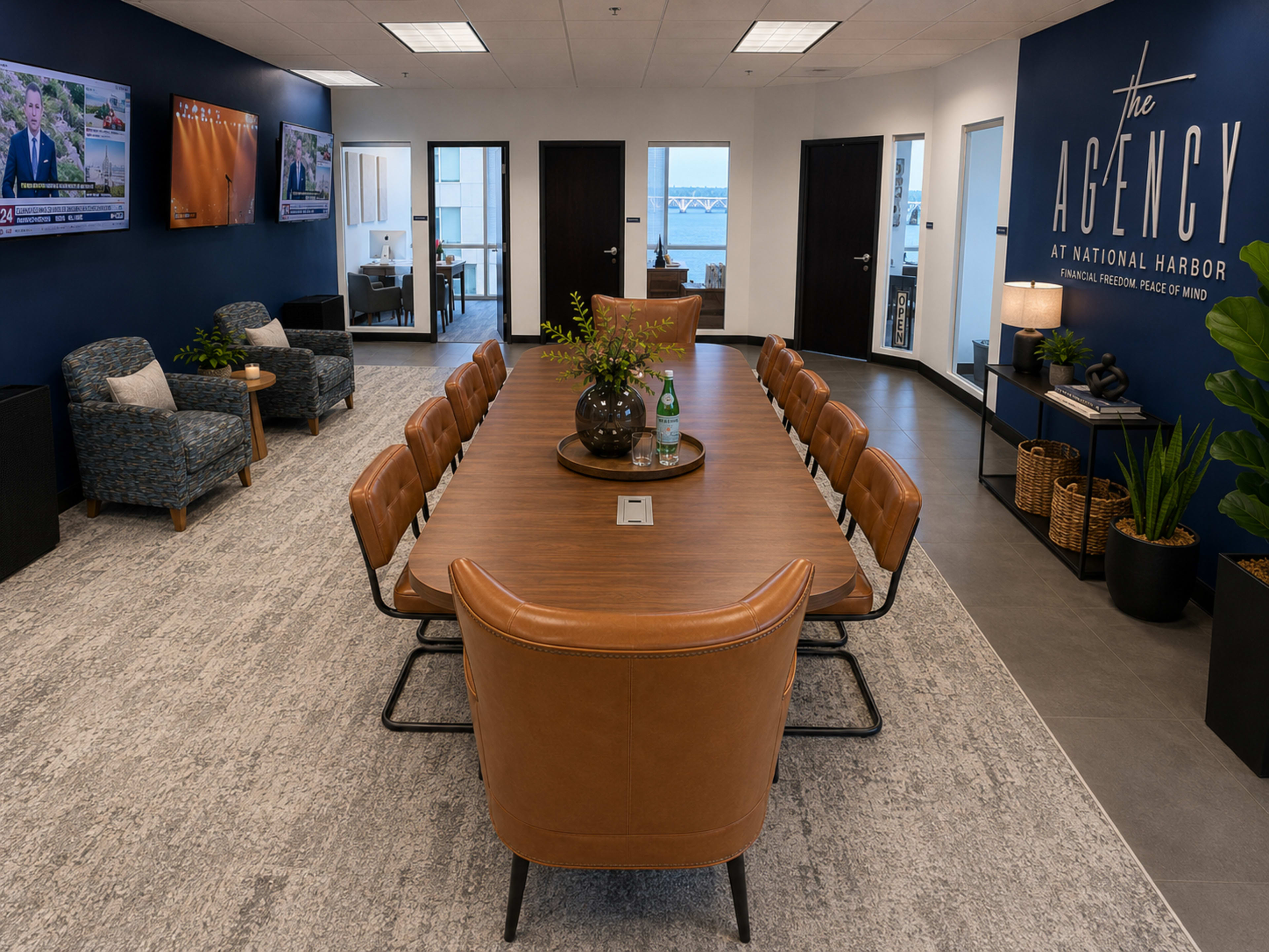 The image shows a modern conference room with a long wooden table surrounded by brown leather chairs, set in a well-lit office space featuring television screens and contemporary decor.